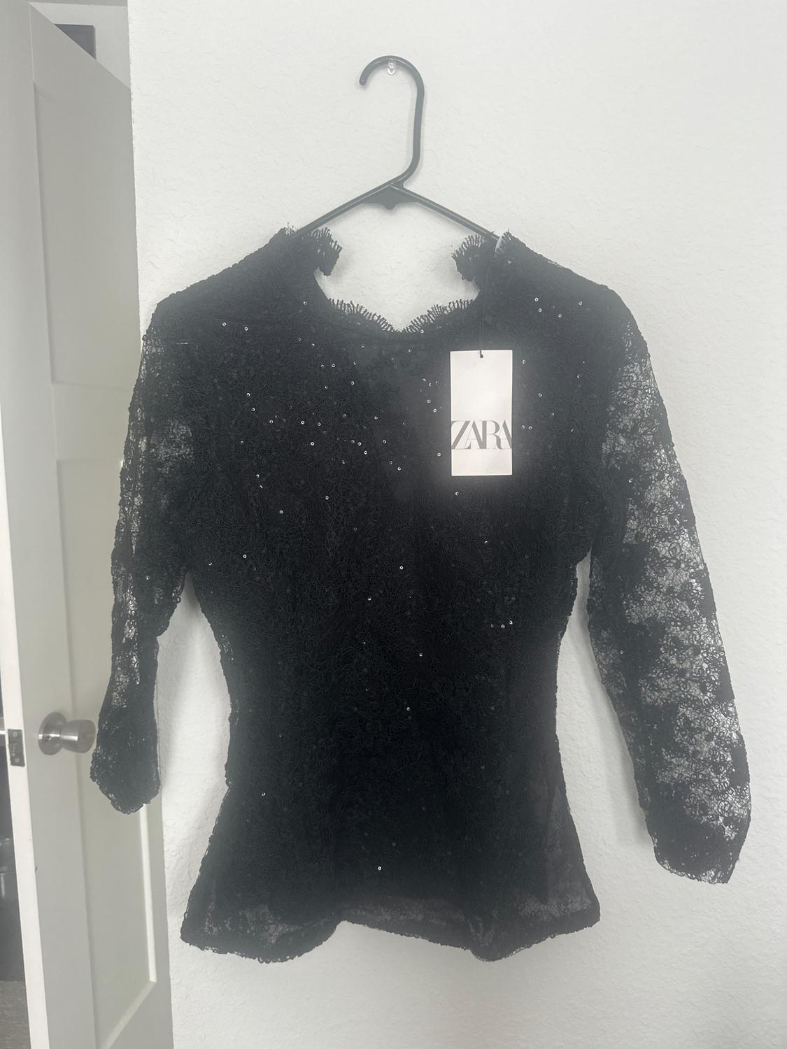 Black sequined lace blouse hanging on a hanger. Visible label with the text "ZARA". White background.