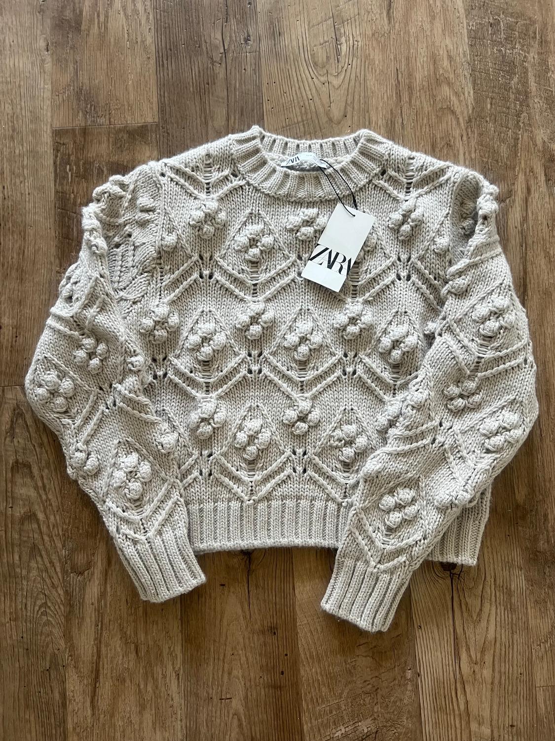 Beige knit sweater with diamond and floral embossed design on a wooden floor. Visible label with text "ZARA".
