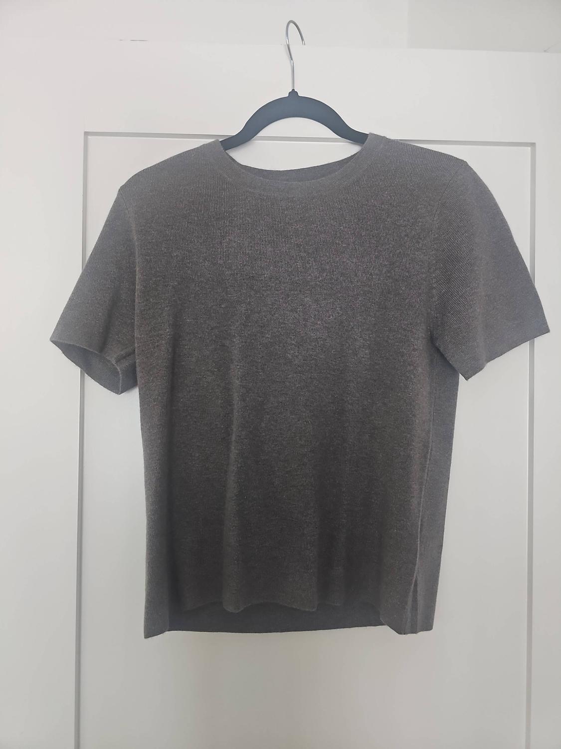 Dark grey short-sleeved knitted T-shirt hanging on a black hanger in front of a white door.
