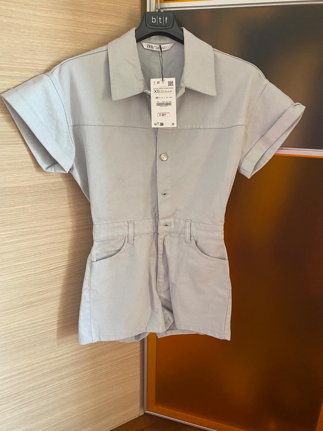 Light grey short-sleeved jumpsuit with a classic collar, hanging on a black hanger. Visible price tag of €39.95.