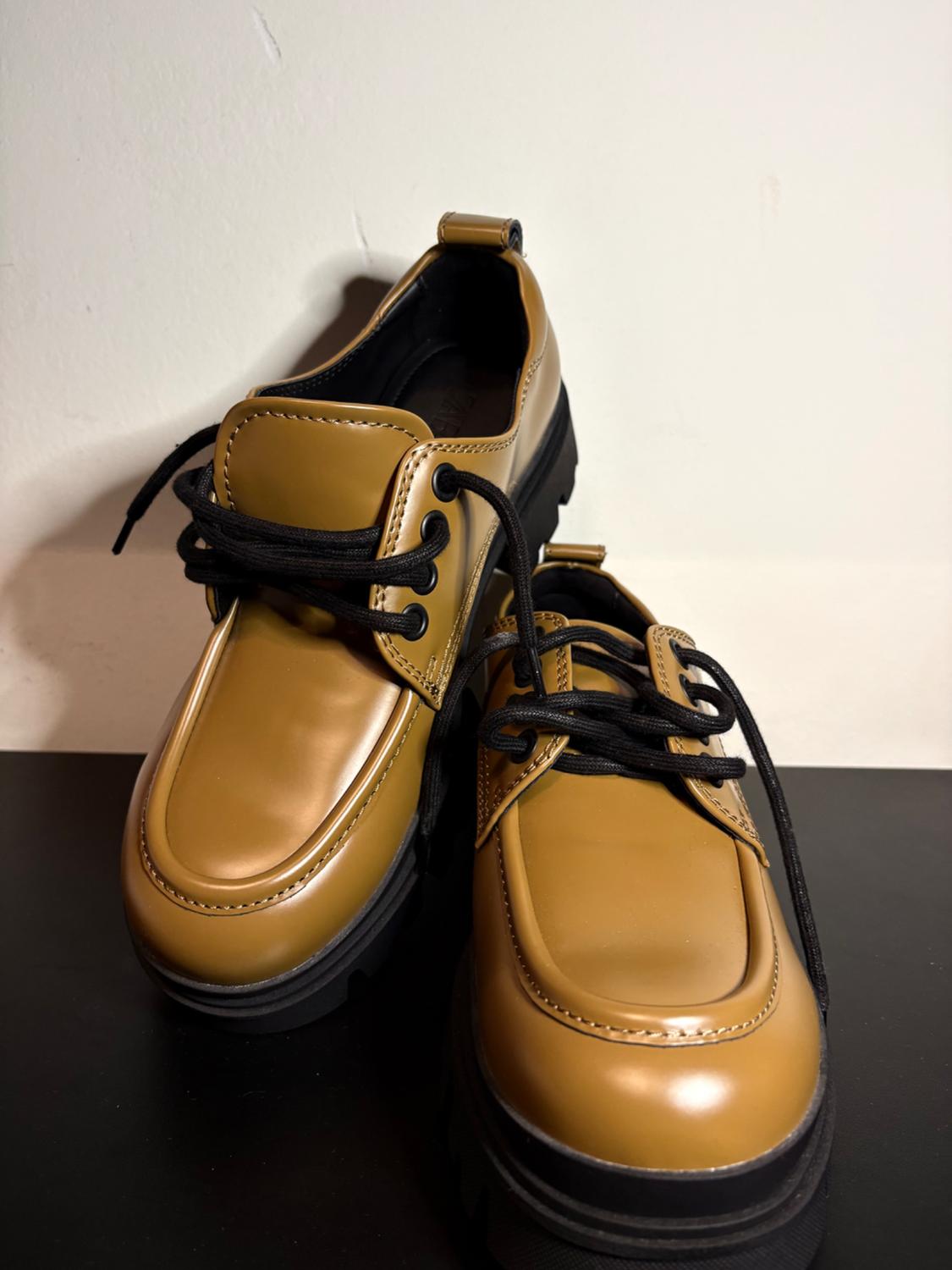 Brown patent leather shoes with black laces and a thick black sole, on a white background and black surface.