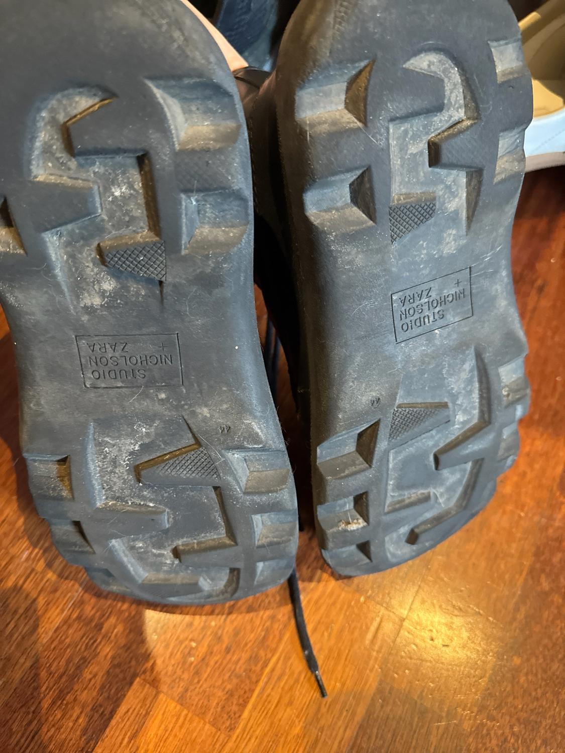 Black boot soles on a wooden floor. They have a ridged tread and the text “STUDIO NICHOLSON + ZARA” alongside the number “44”.