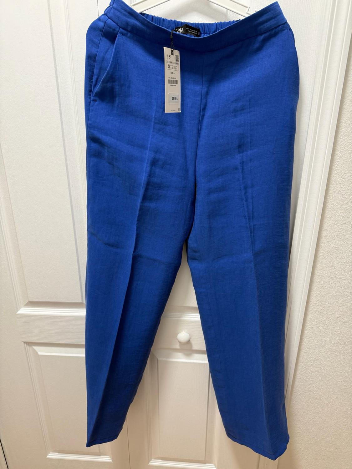 Blue pants hanging on a hanger over a white door. They feature an elastic waistband, a front crease, and a tag with the text 49.90.