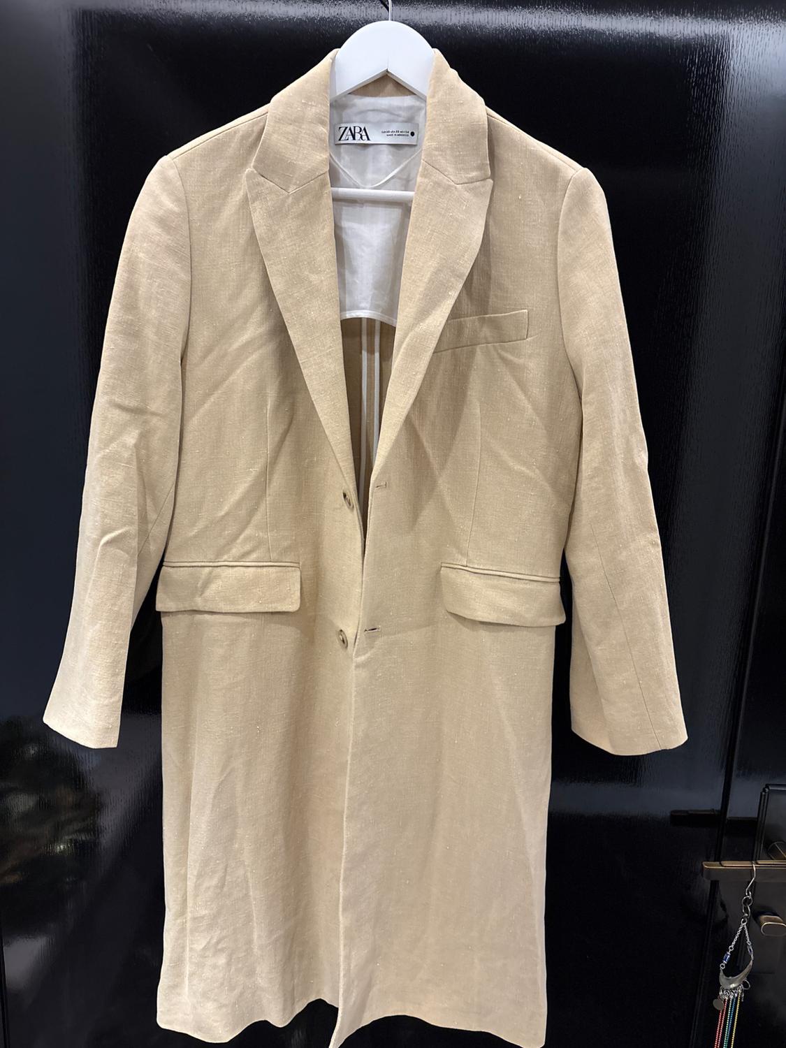 Beige linen coat hanging on a white hanger with a Zara label, against a shiny black background.