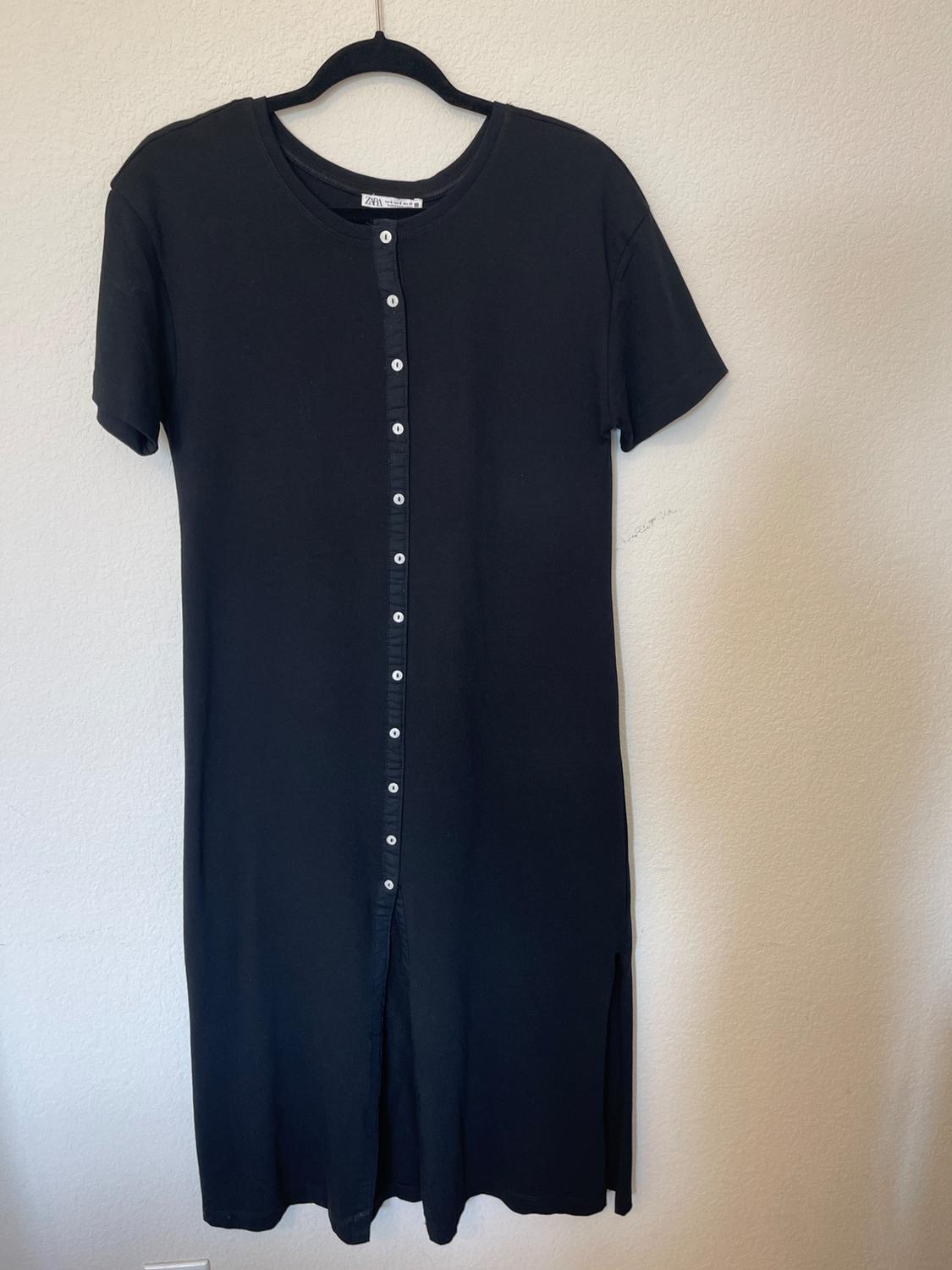 Black short-sleeved dress with white buttons down the front, hanging on a hanger against a white wall.