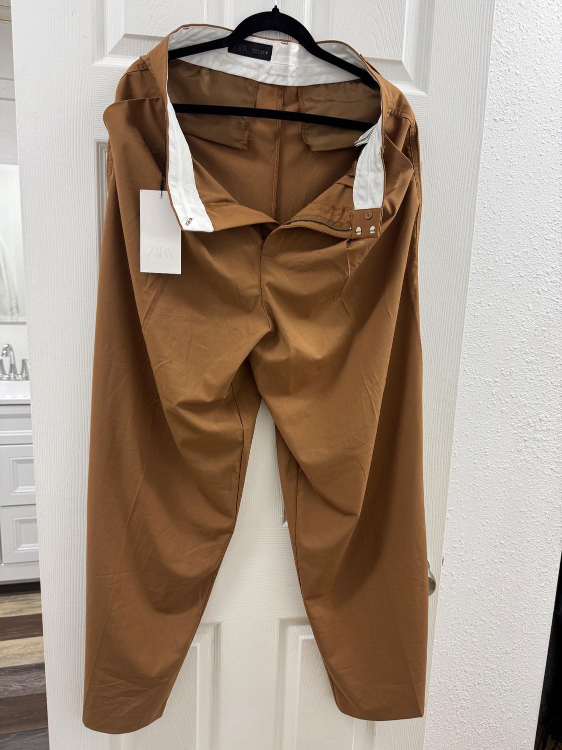 Brown trousers hanging on a black hanger over a white door. Visible label with text "ZARA". Partially visible bathroom background.