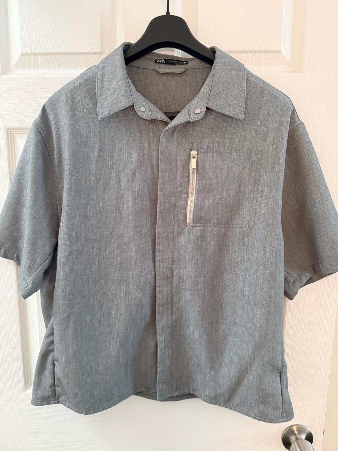 Gray short-sleeved shirt with a zippered pocket, hanging on a black hanger in front of a white door. ZARA label visible.