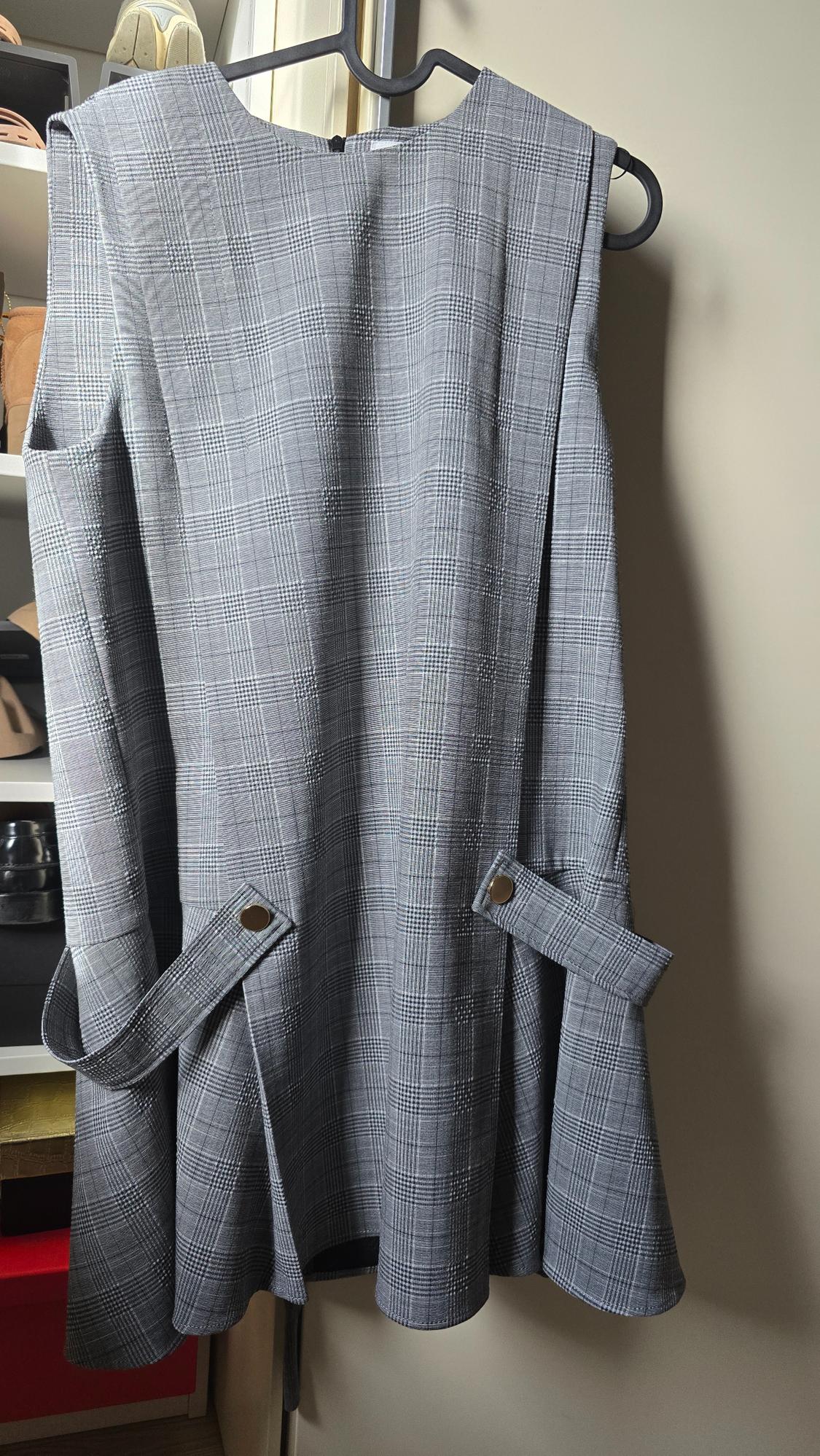 Grey dress with a checked print, sleeveless, hanging on a black hanger. Background with visible shelves and shoes.