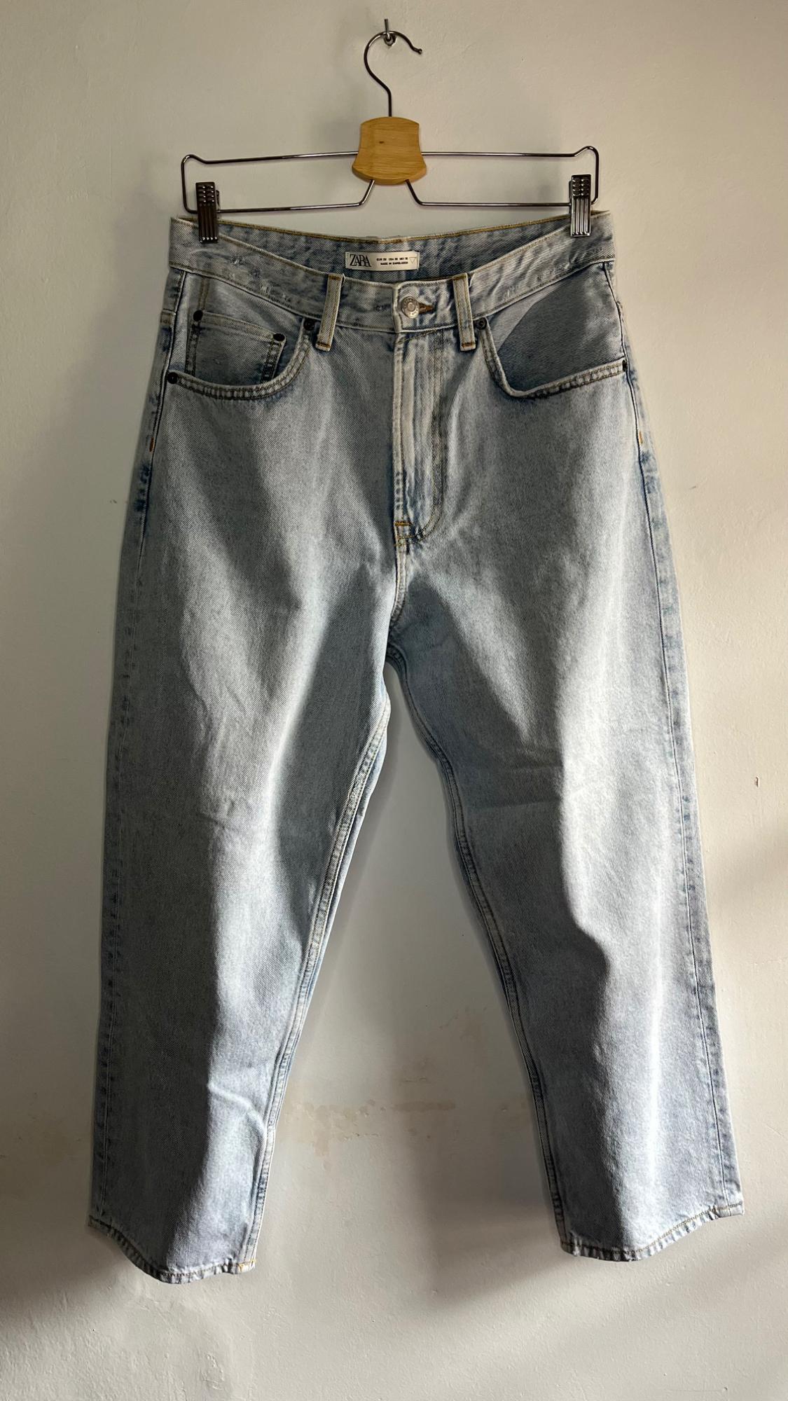 Light blue jeans hanging on a wooden hanger against a white wall. Label visible on the waistband.