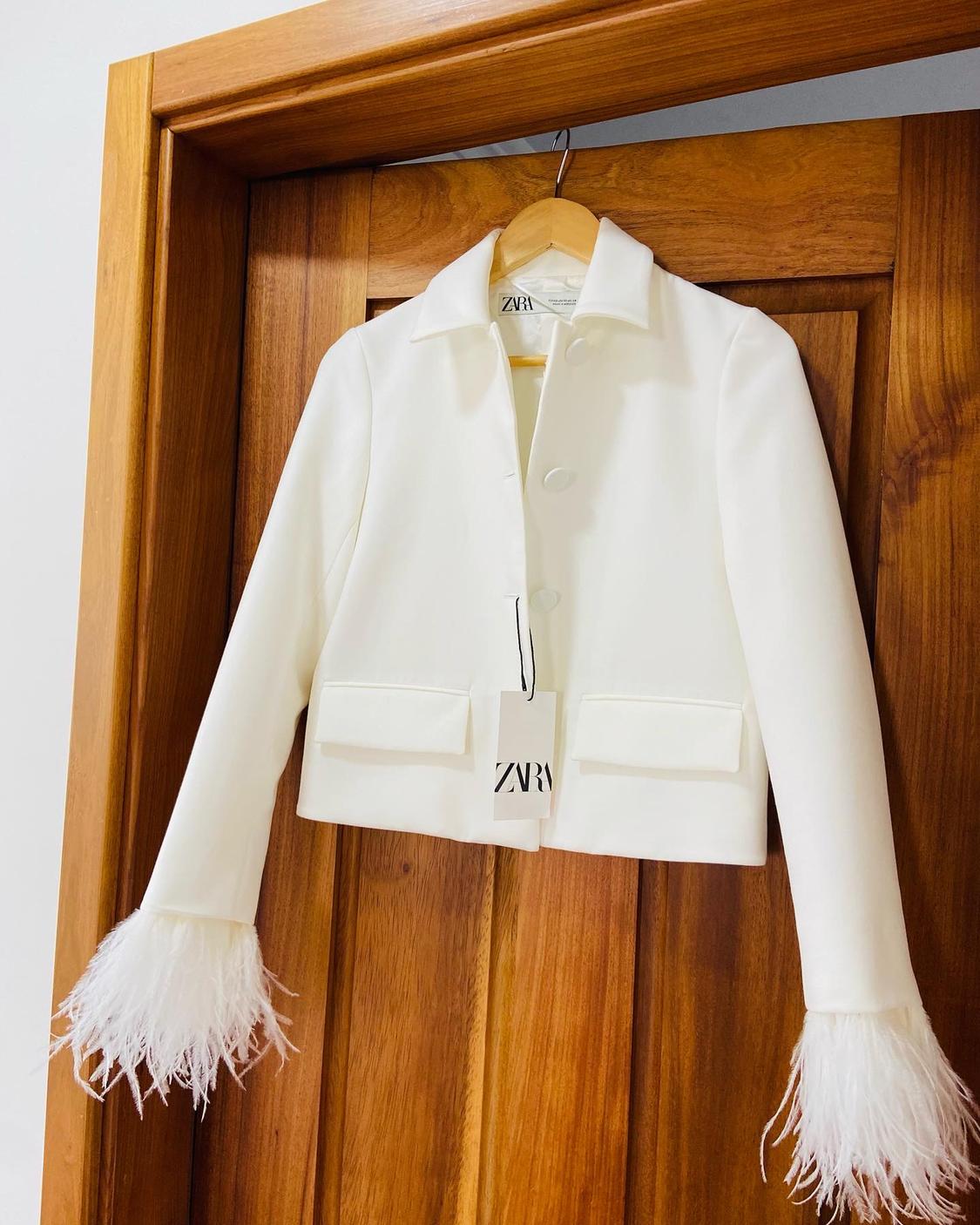 Front view of a white jacket with buttons and front flap pockets. Cuffs decorated with feathers.