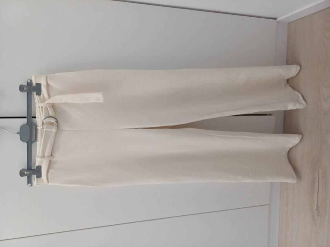 White trousers hanging on a grey hanger against a white wall and light wood floor.