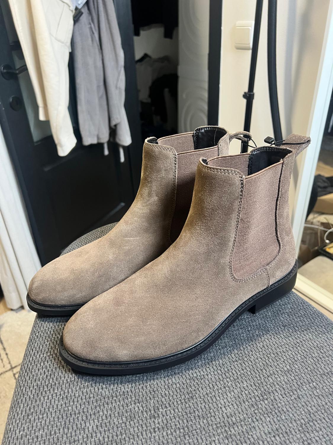 Light brown suede boots on a grey surface, with a black door and hanging clothes in the background.