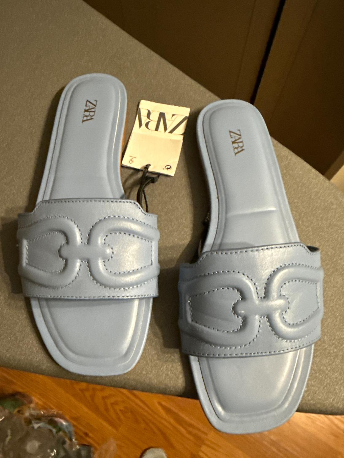Blue flat sandals with a decorative design on the top. Zara tag visible on a gray surface.