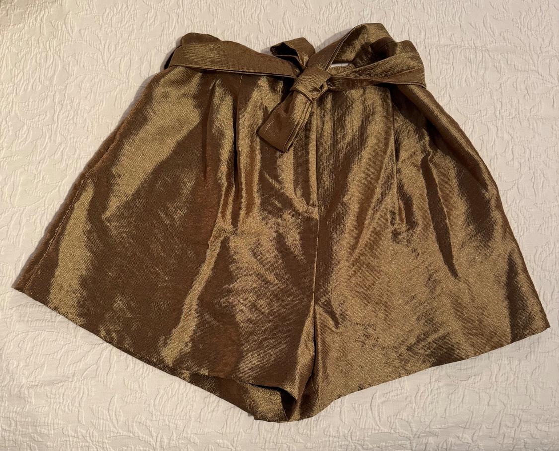 Shiny gold shorts with a knotted belt, on a textured white bedspread.