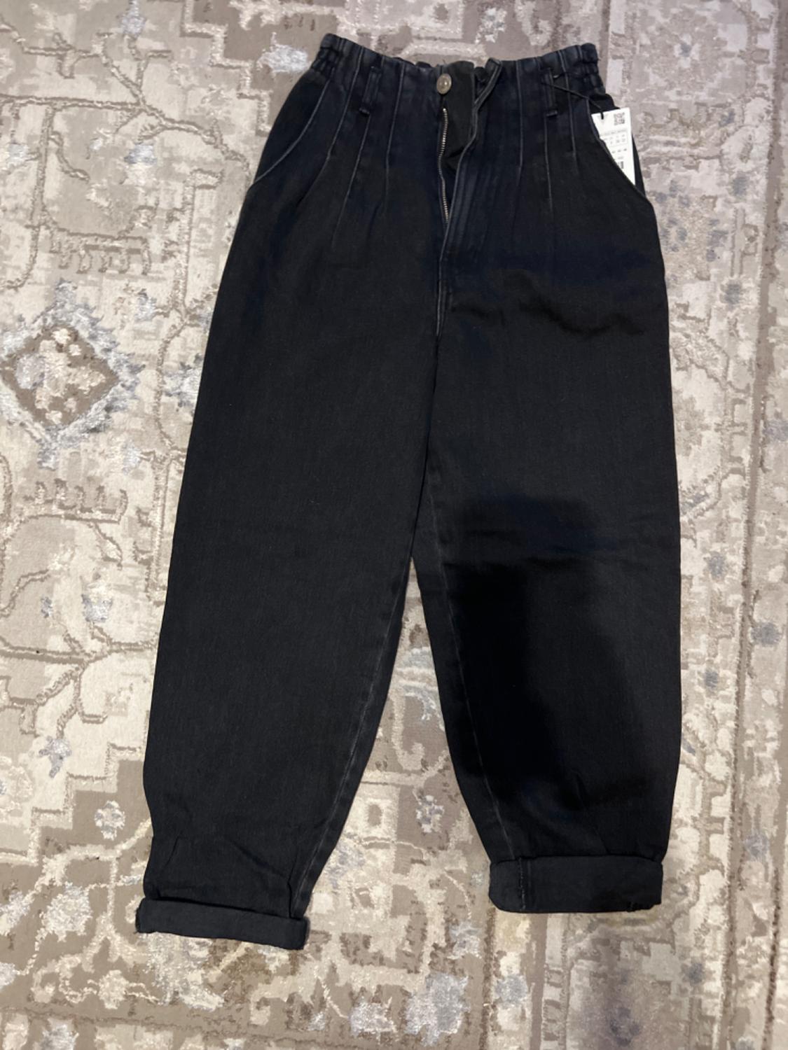 Black denim pants with pleats, elastic waistband, and rolled hem, on a beige rug with a geometric design. Visible tag.