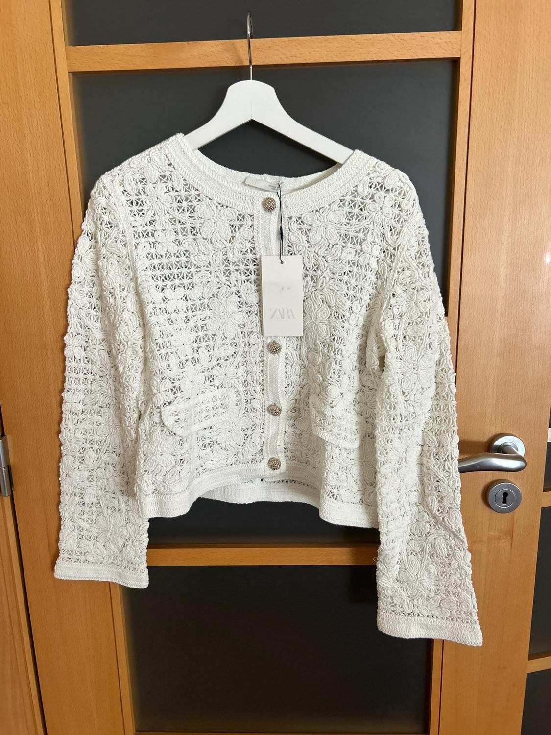 White open-knit jacket with buttons and pockets, hanging on a coat hanger over a wooden door. Label bearing the text ZARA.