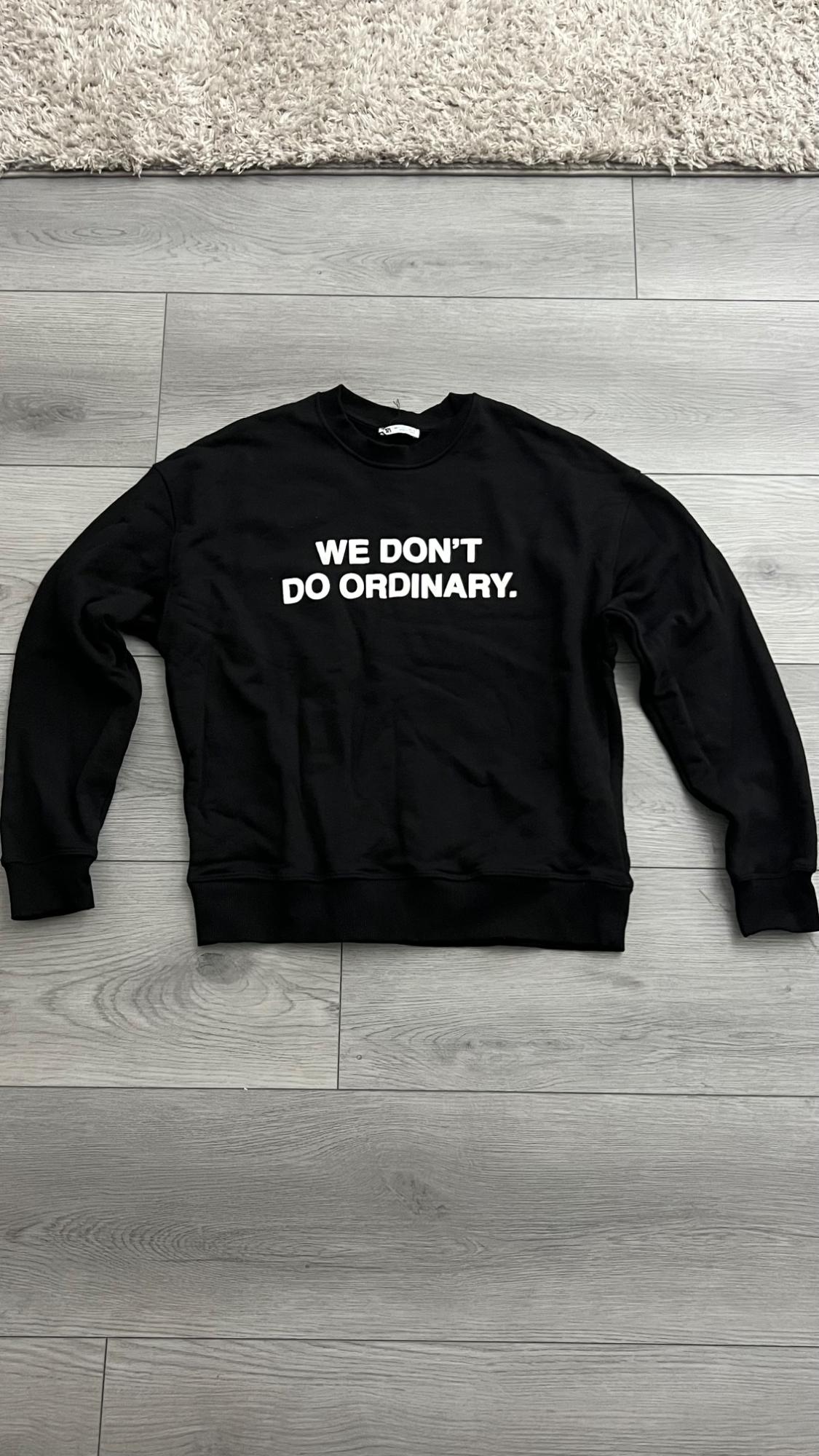 Casual black sweatshirt on a gray wooden floor with the text WE DON'T DO ORDINARY. The edge of a white rug is visible at the top.