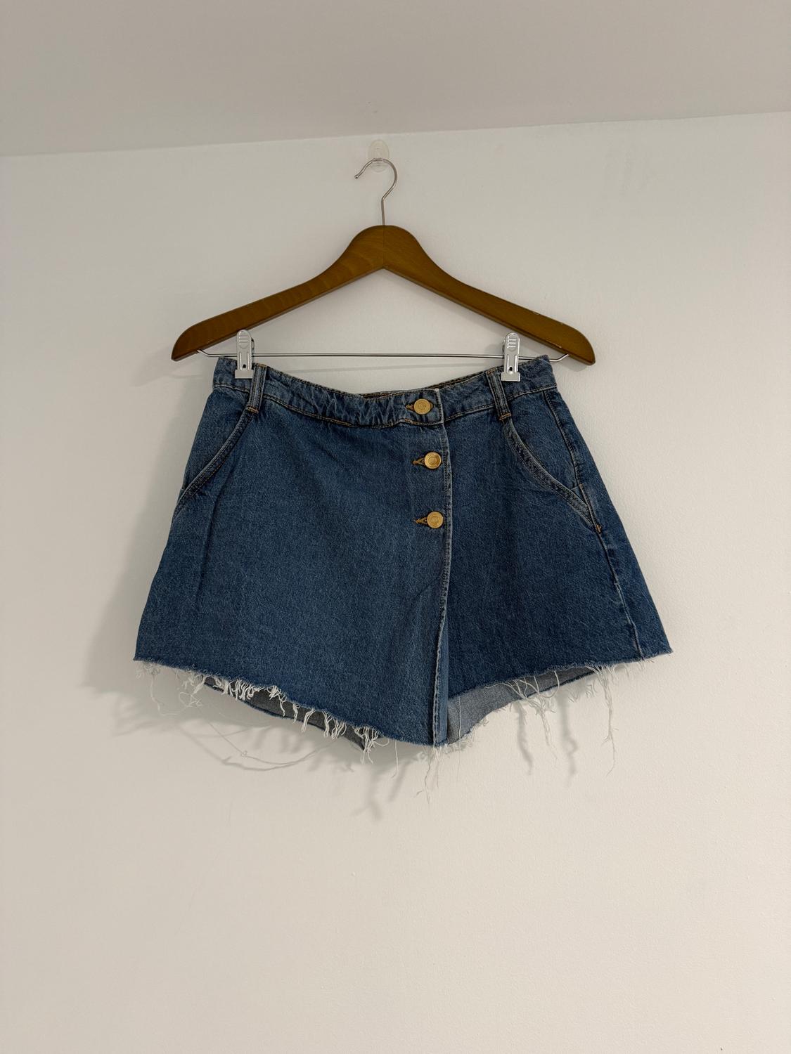 Blue denim mini skirt with gold buttons and a frayed hem, hanging on a wooden hanger against a white wall.