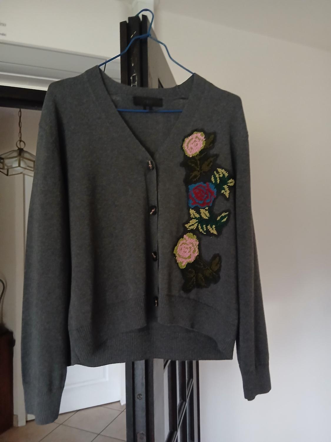 Grey cardigan hanging on a blue hanger, with pink and blue floral embroidery on the right side, interior background of a house.
