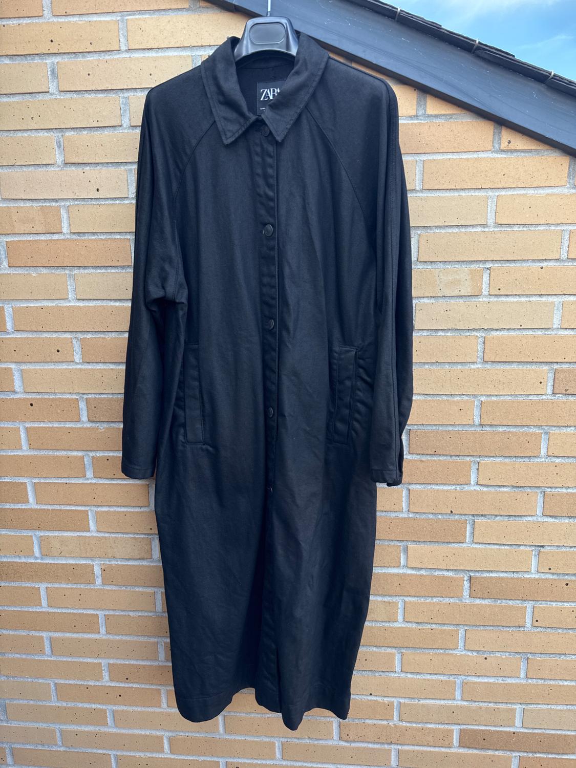 Black coat hanging on a hanger in front of a brick wall. Visible label with the text "ZARA".