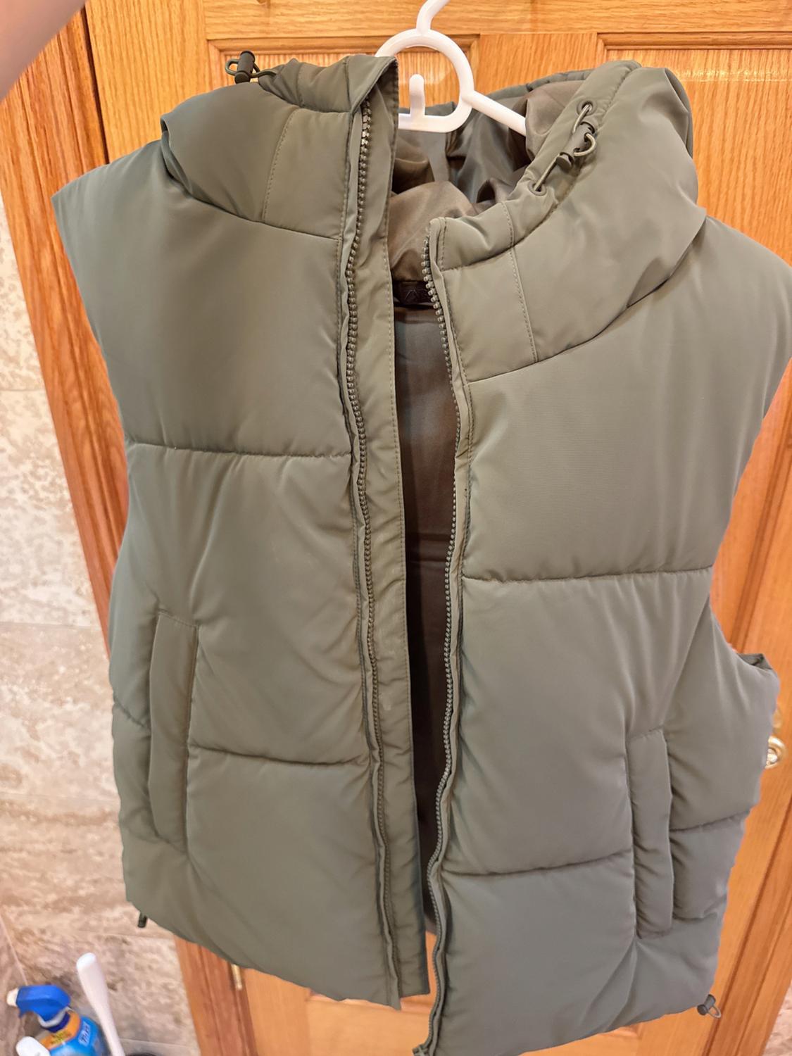Olive green padded gilet hanging on a white hanger, with the zip open, over a light wooden door.