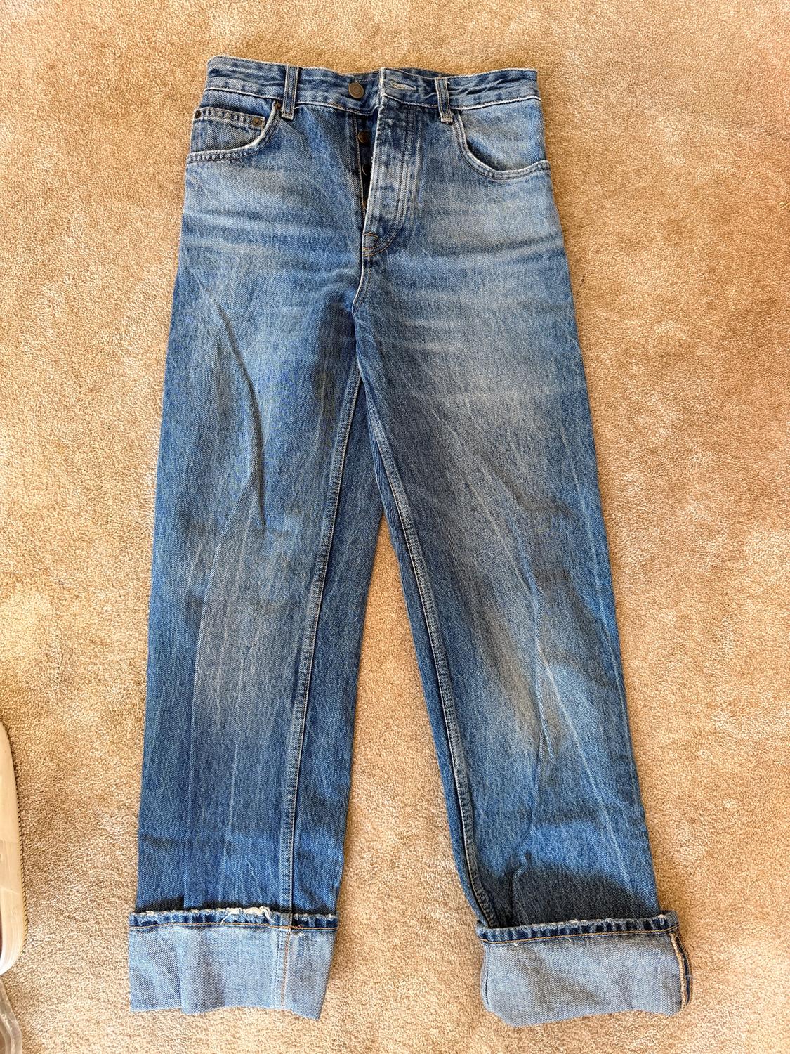Blue denim jeans with rolled hem, placed on a beige rug. Part of a trainer visible on the left.