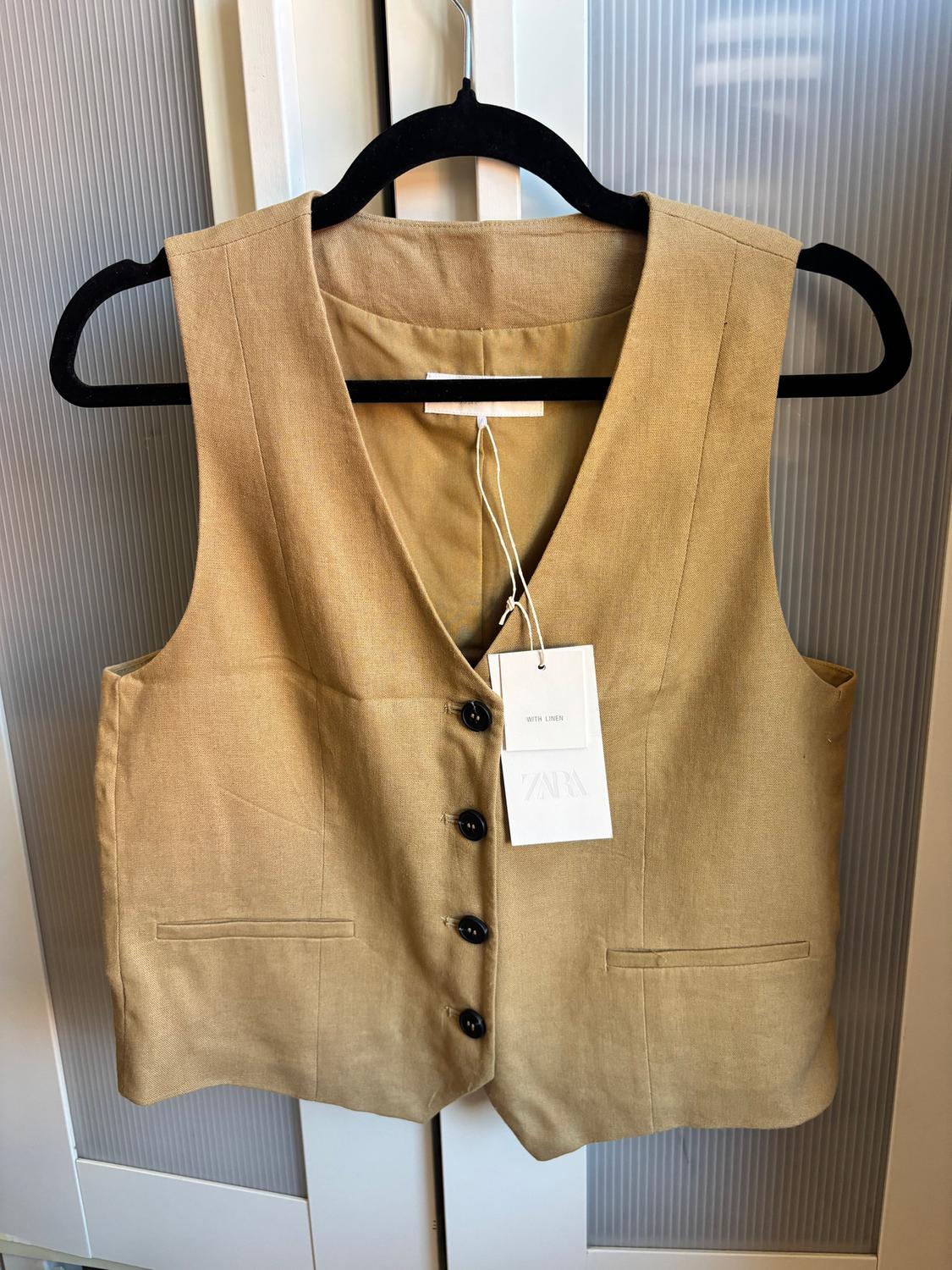 Beige waistcoat hanging on a black hanger, with dark buttons and a white label reading "WITH LINEN ZARA", against a background of white doors.