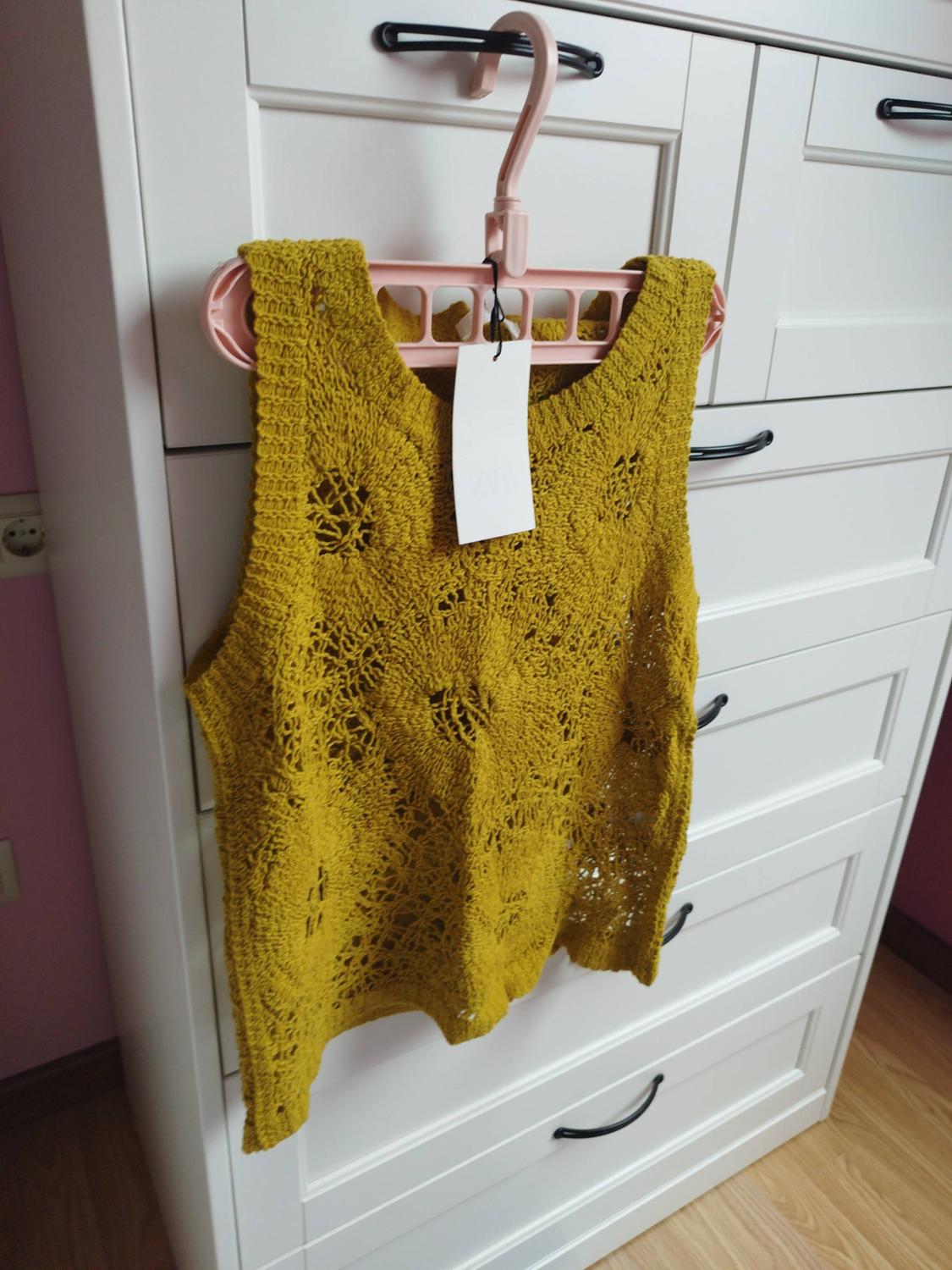 Top de punto calado sin mangas color mostaza con etiqueta ZARA, colgado en una percha rosa de una cómoda blanca en un ambiente hogareño.