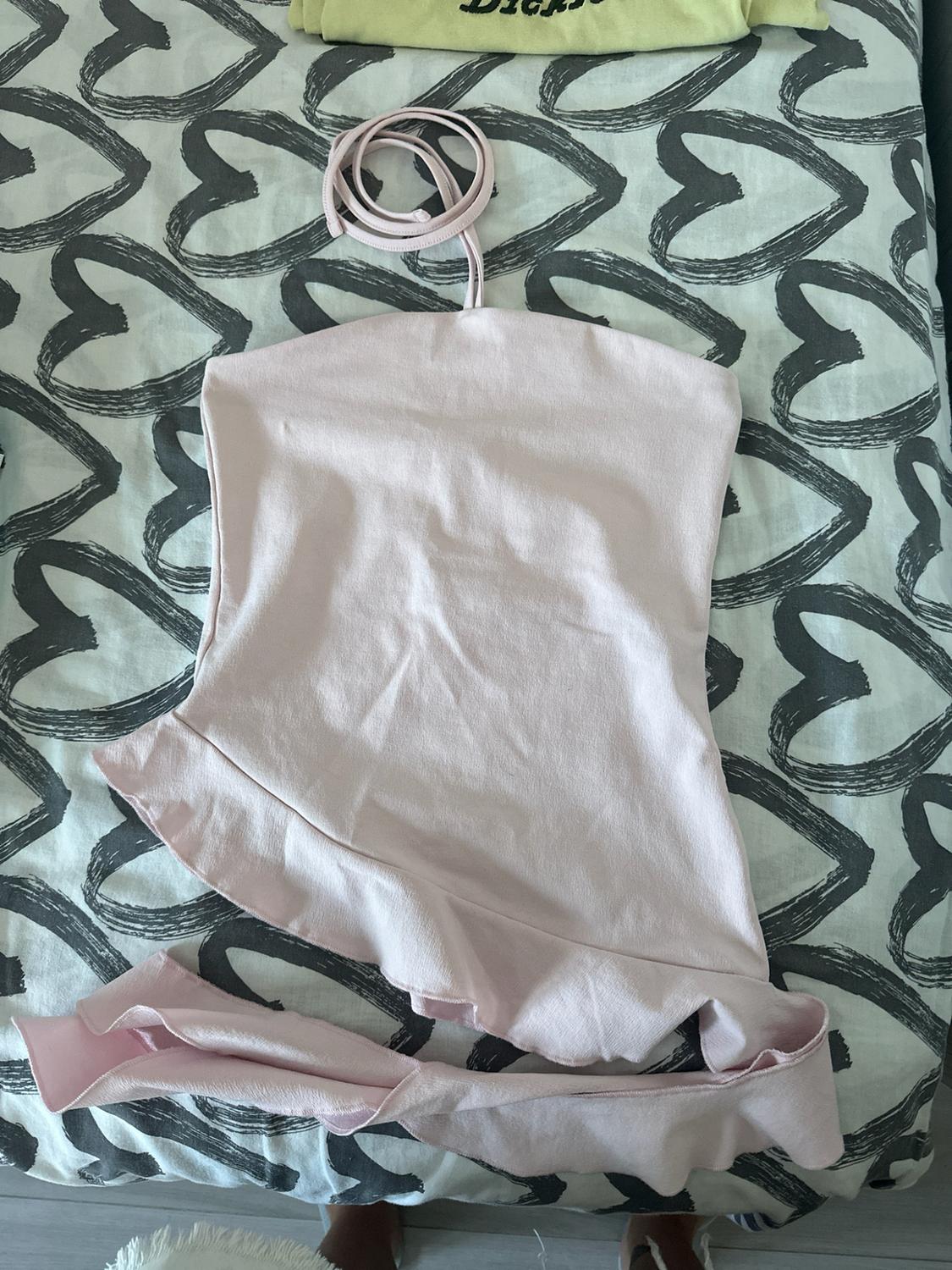 Light pink halter-neck top with an asymmetrical ruffled hem on a heart-print sheet. A yellow garment with text peeks out from the top.