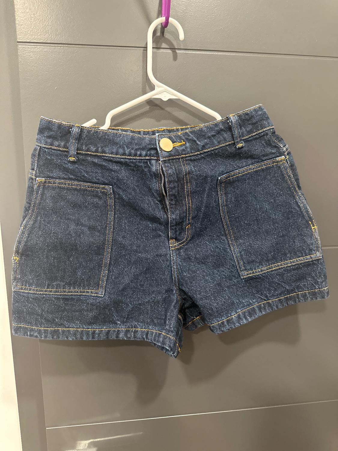 Blue denim shorts with large pockets, hanging on a white hanger over a grey door.
