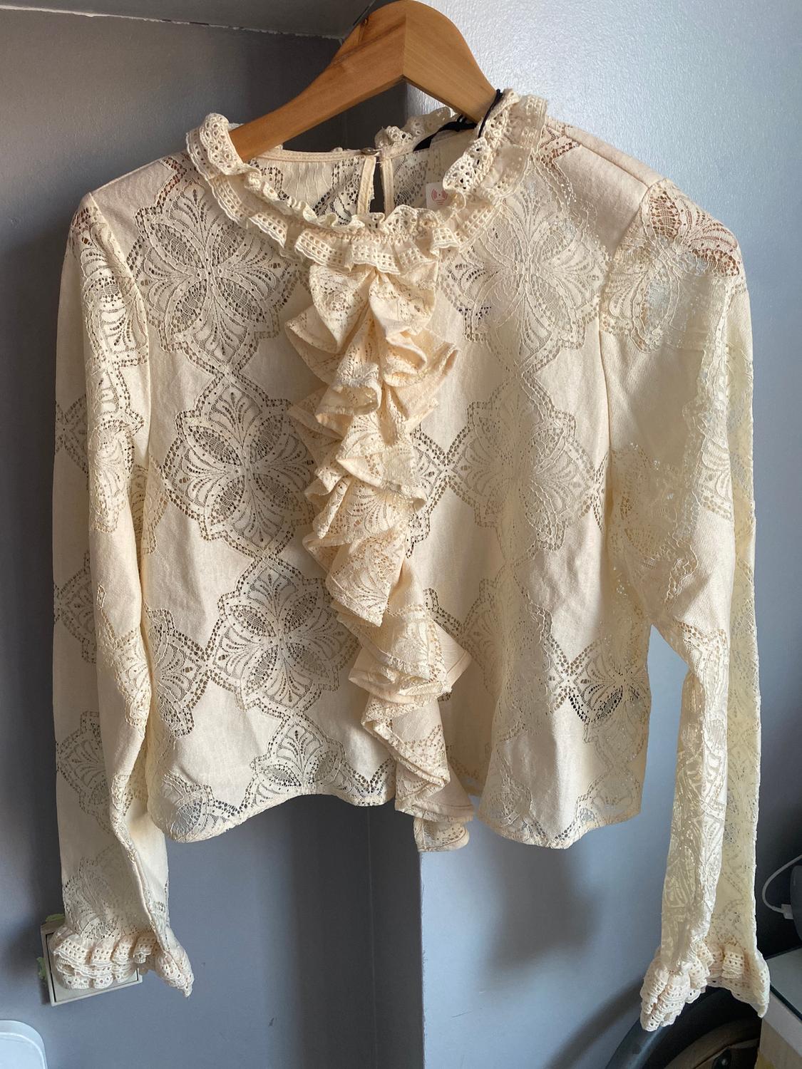 Beige lace blouse with front ruffles, hanging on a wooden hanger against a grey wall.