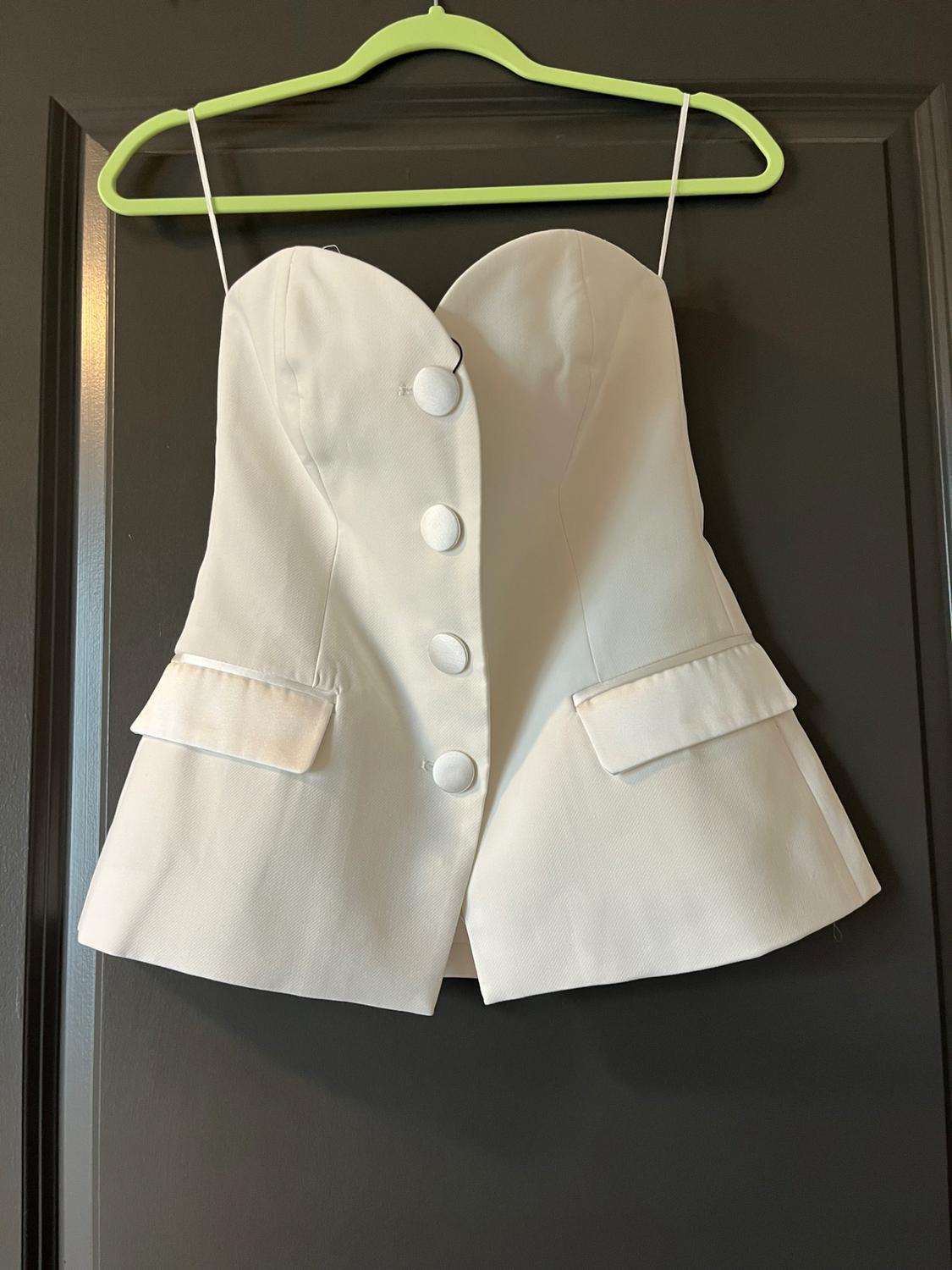 White strapless sweetheart neck top with buttons and flap pockets, hanging on a light green hanger over a dark door.