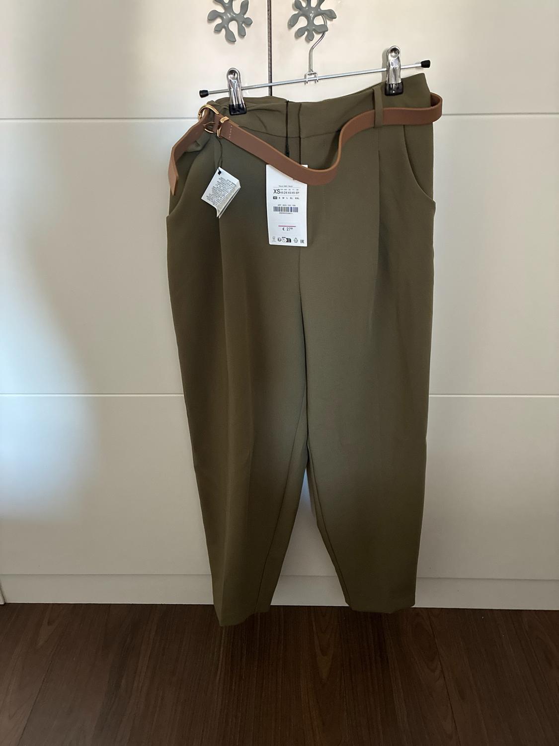 Olive-green trousers with a brown belt hanging in a white wardrobe. Visible label with the text "XS" and "€ 27".