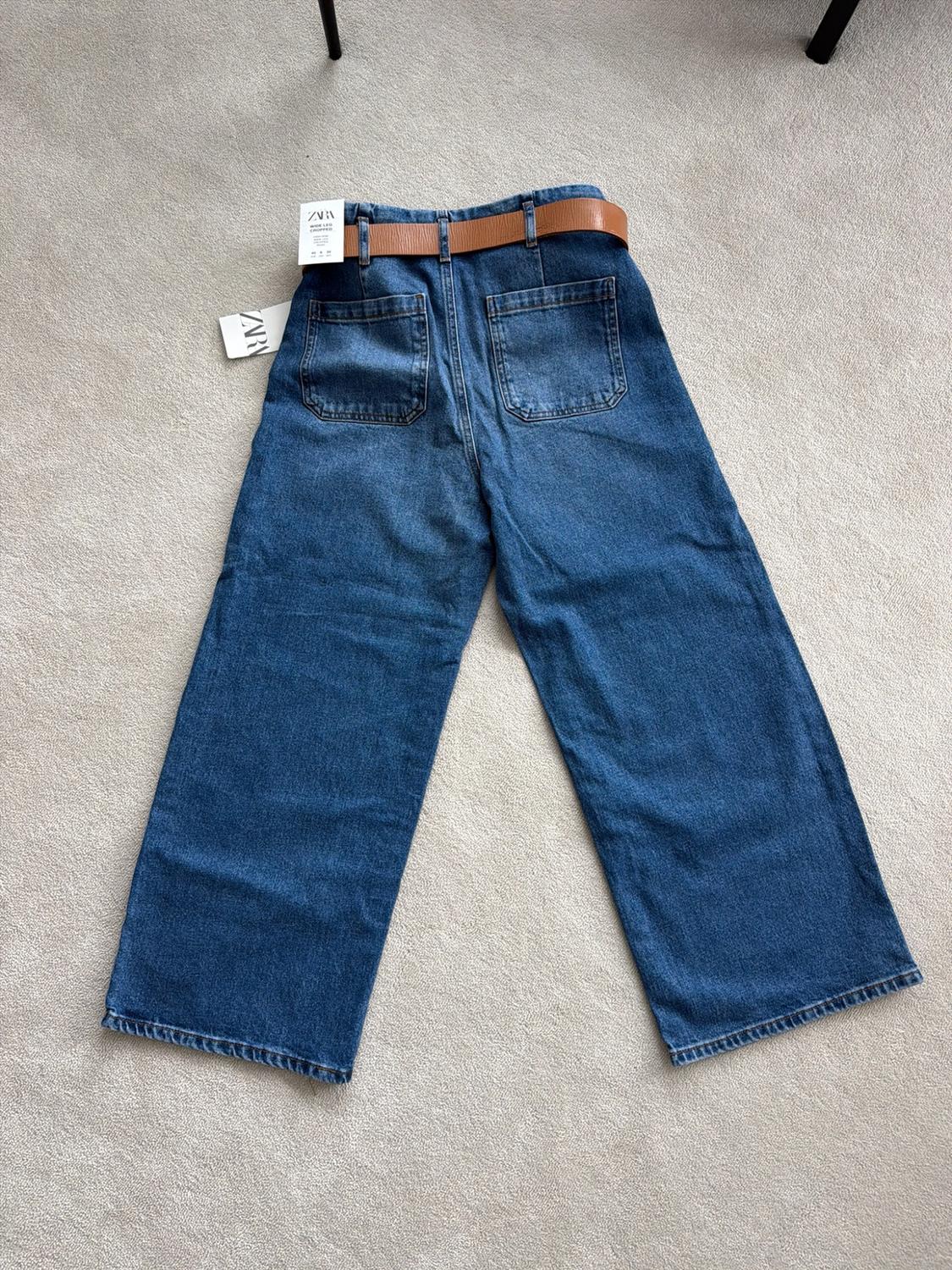 Blue wide-leg denim jeans with a brown belt and ZARA labels, seen from behind on a rug.