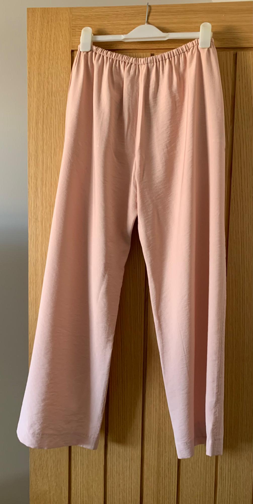 Pale pink trousers hanging on a white hanger over a wooden door. Soft fabric and wide-leg design.