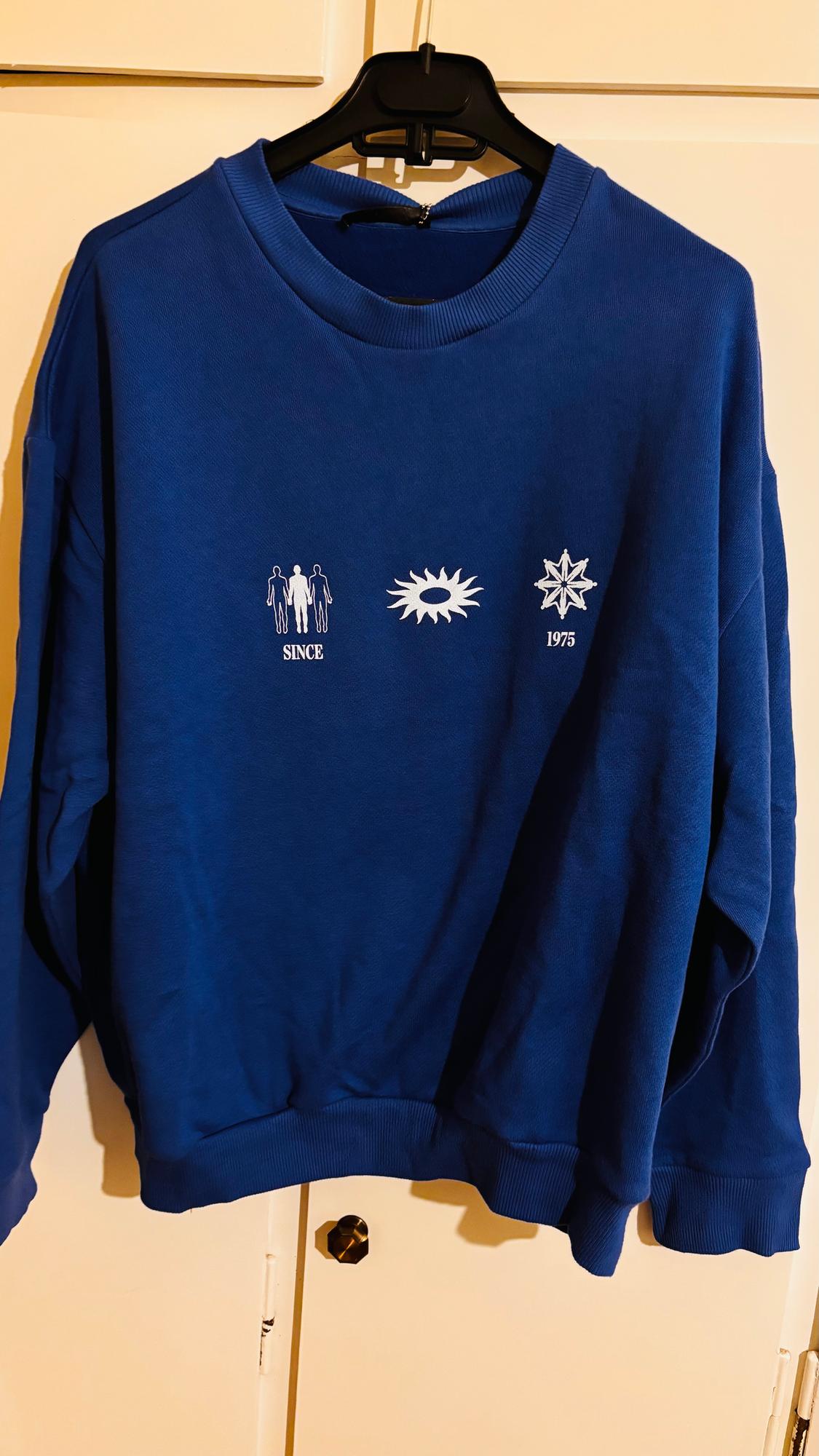 Blue sweatshirt hanging on a hanger, with white graphics of human figures, a sun, and a star. Text: "SINCE 1975".
