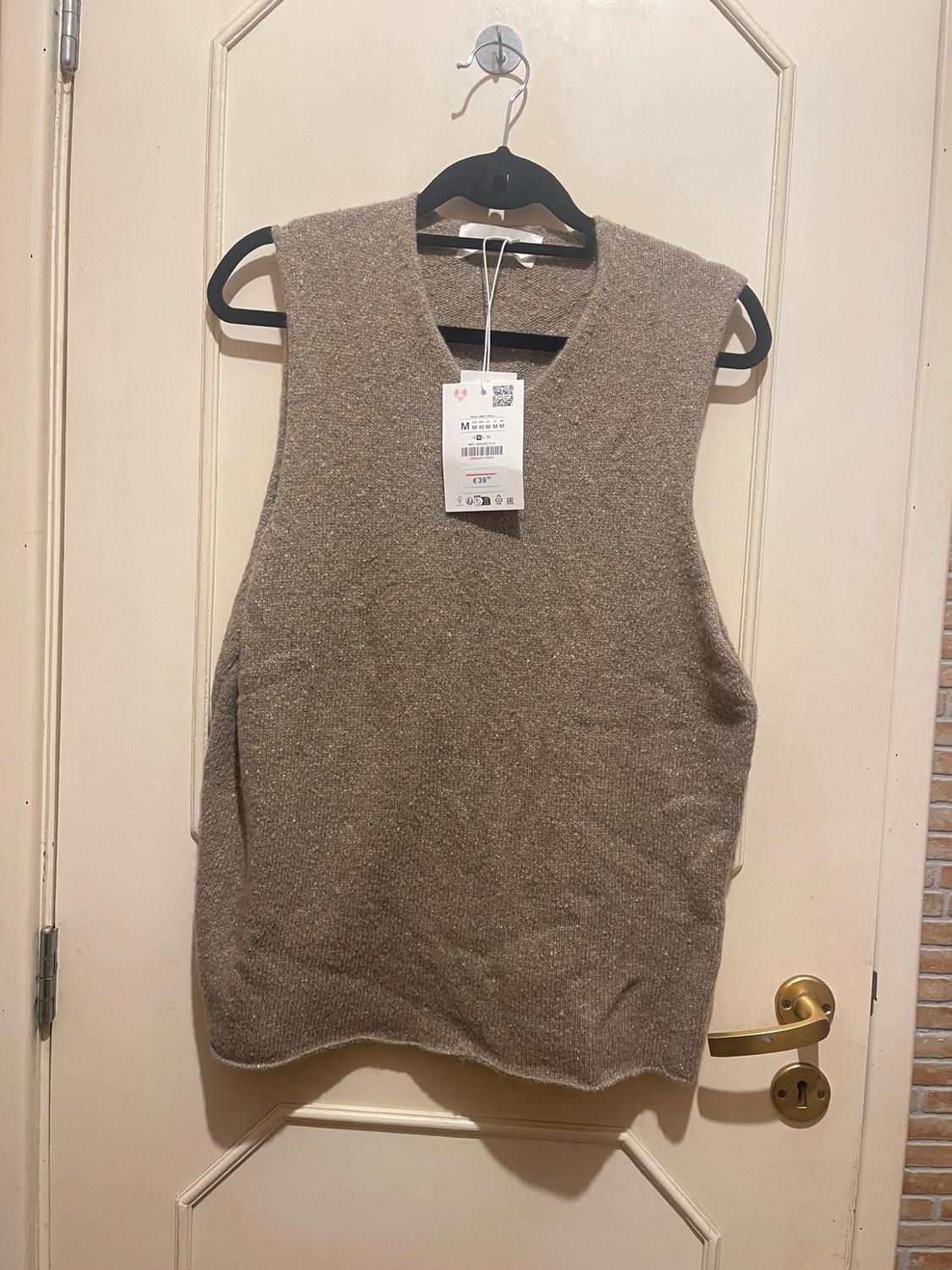 Beige knitted waistcoat with a V-neck hanging on a black hanger over a white door. Tag with the text M and €39.