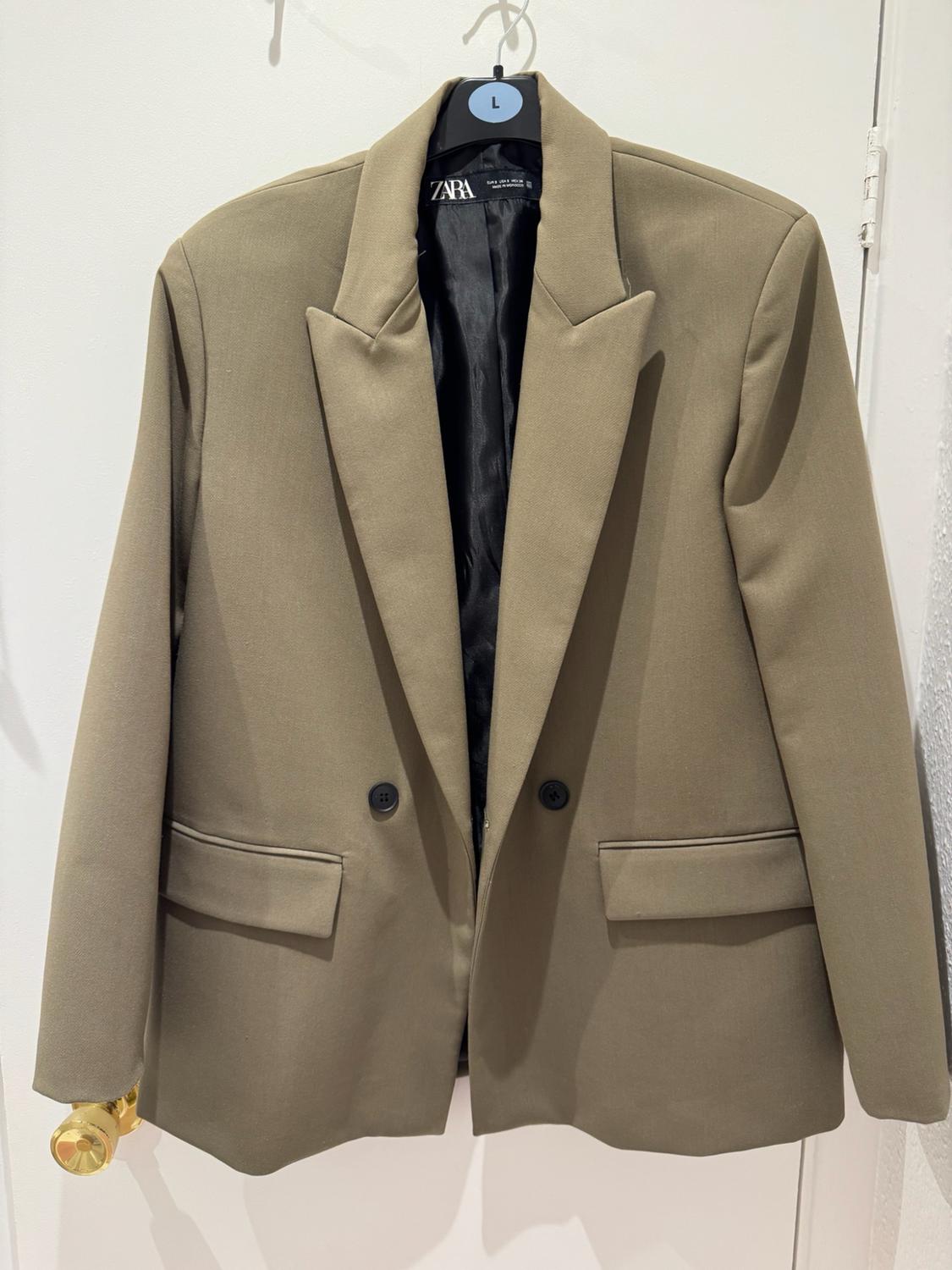 Beige blazer hanging on a white door with a golden handle. It has lapels, buttons and pockets. ZARA label and a hanger with the letter L.
