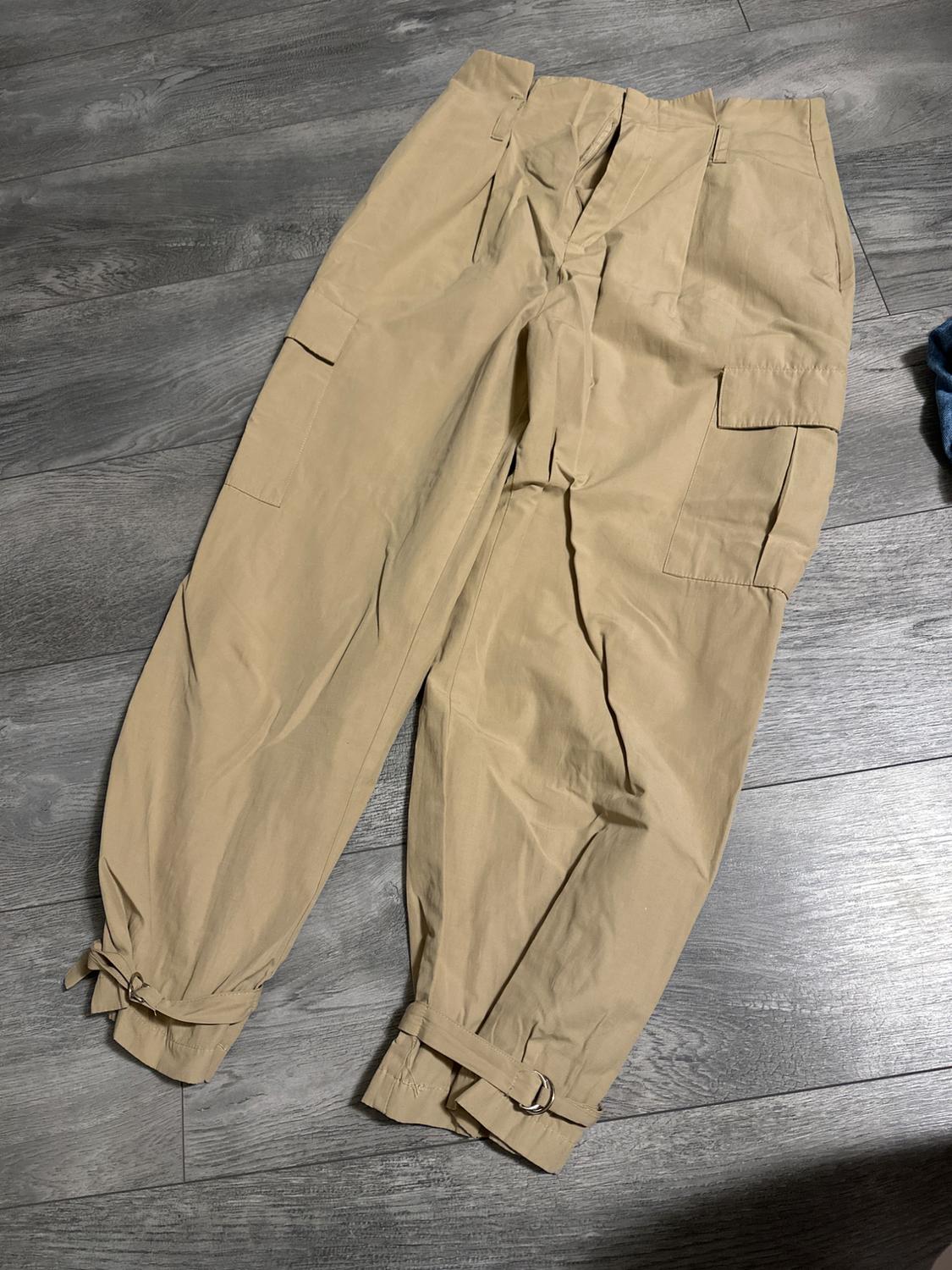 Beige cargo trousers with side pockets and adjustable ankle straps, on a grey wooden floor.