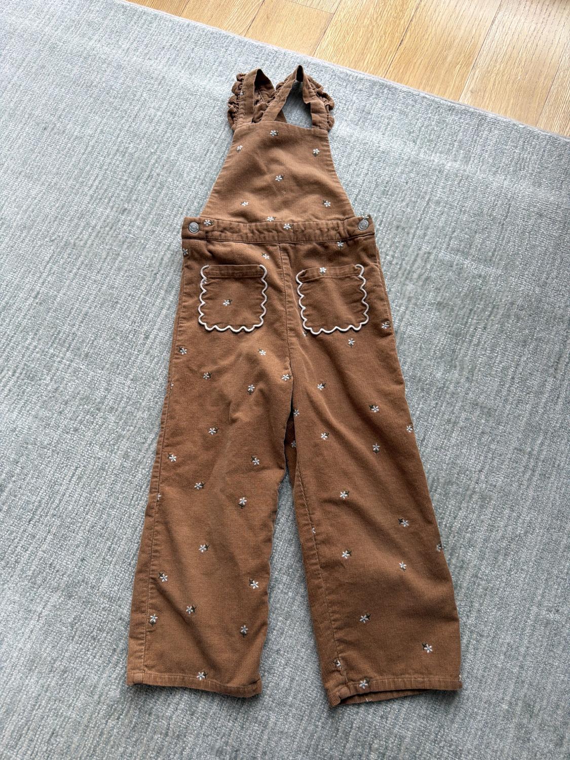 Brown corduroy dungarees with white flowers and wavy piped pockets, laid out on a grey rug in a relaxed setting.