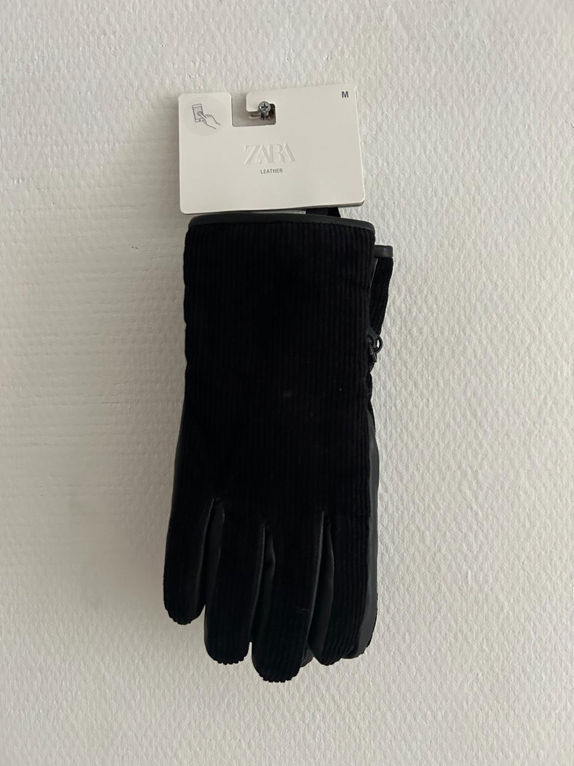 Black corduroy gloves hanging on a white wall with a cardboard tag that reads "ZARA LEATHER" and size "M".