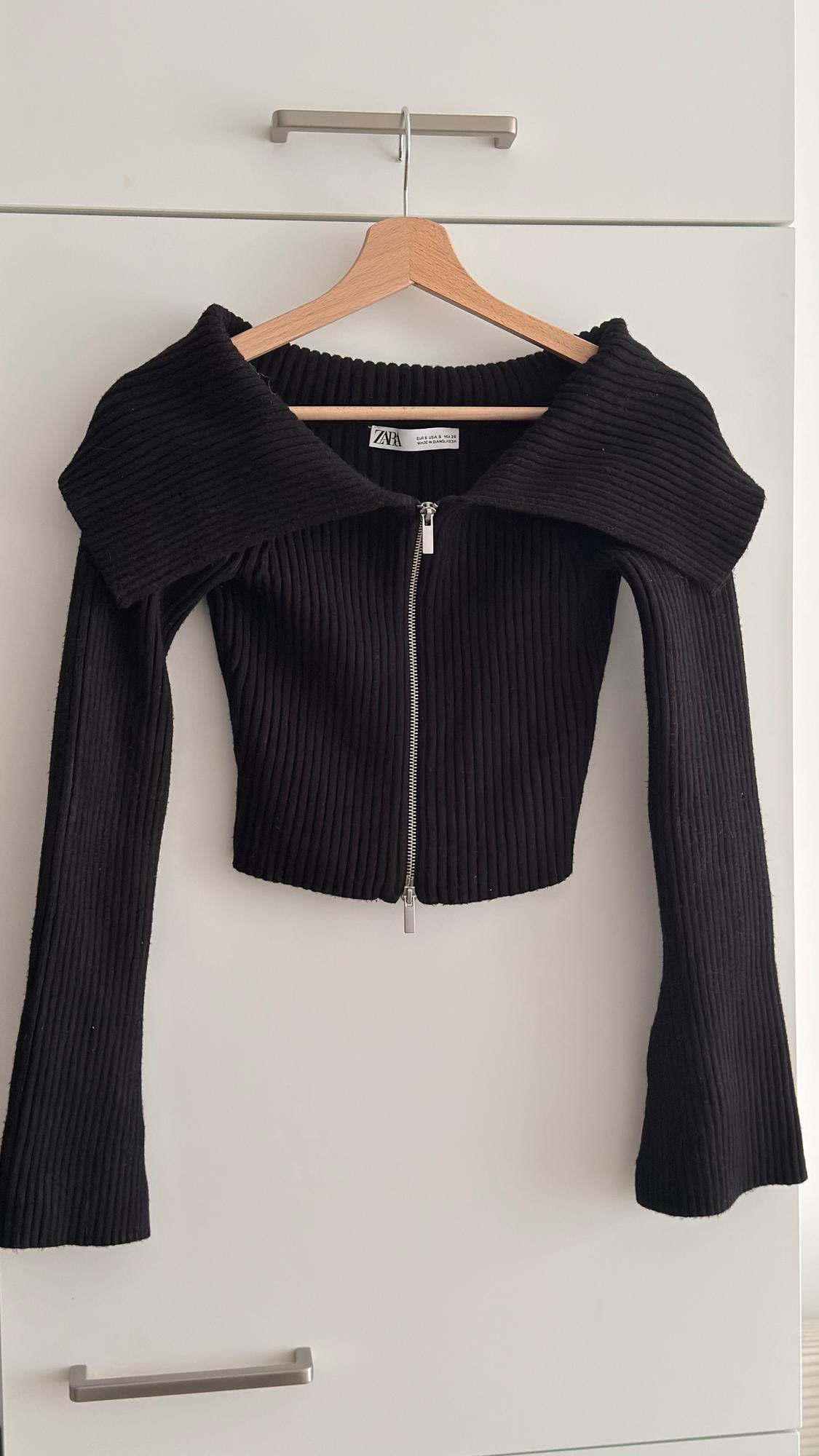 Black ribbed knit jacket with a front zip, hanging on a wooden hanger in front of a white wardrobe.