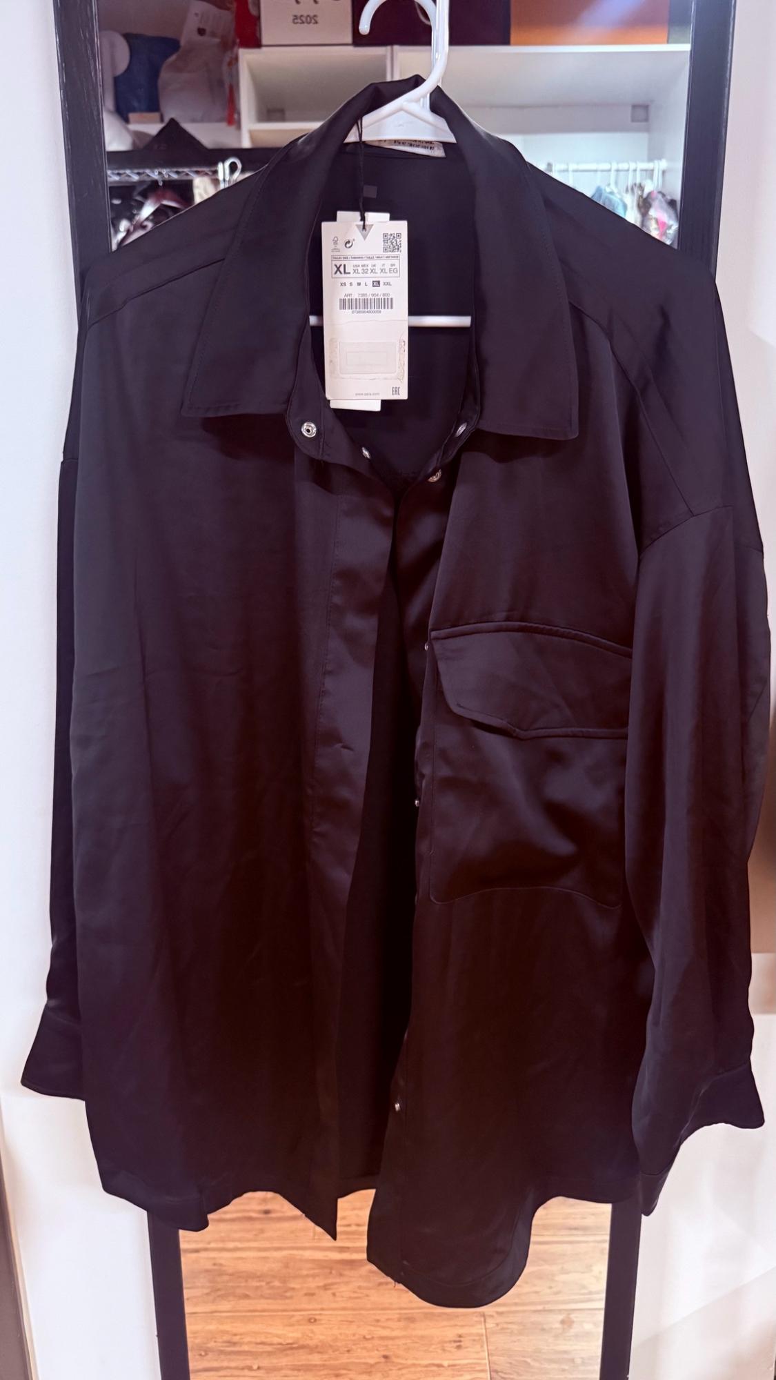 Black long-sleeved button-down shirt with a pocket, hanging on a hanger in front of a mirror. An XL tag is visible.