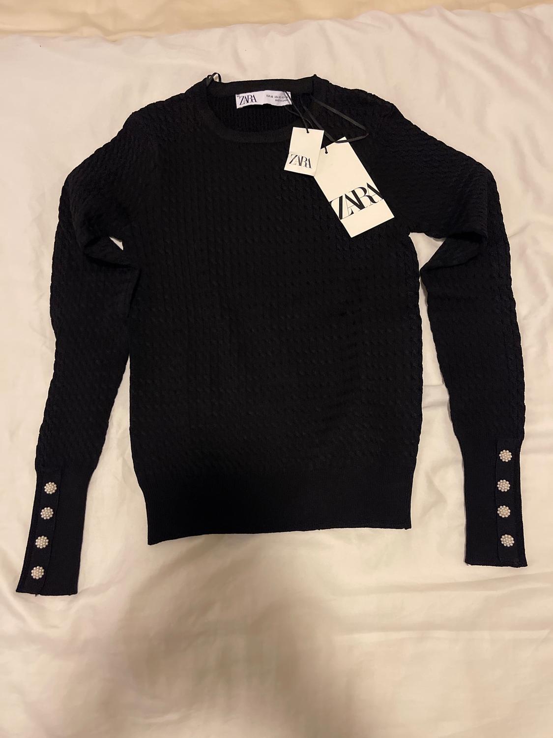 Black long-sleeved textured jumper with decorative buttons on the cuffs and visible brand tags on a white surface.