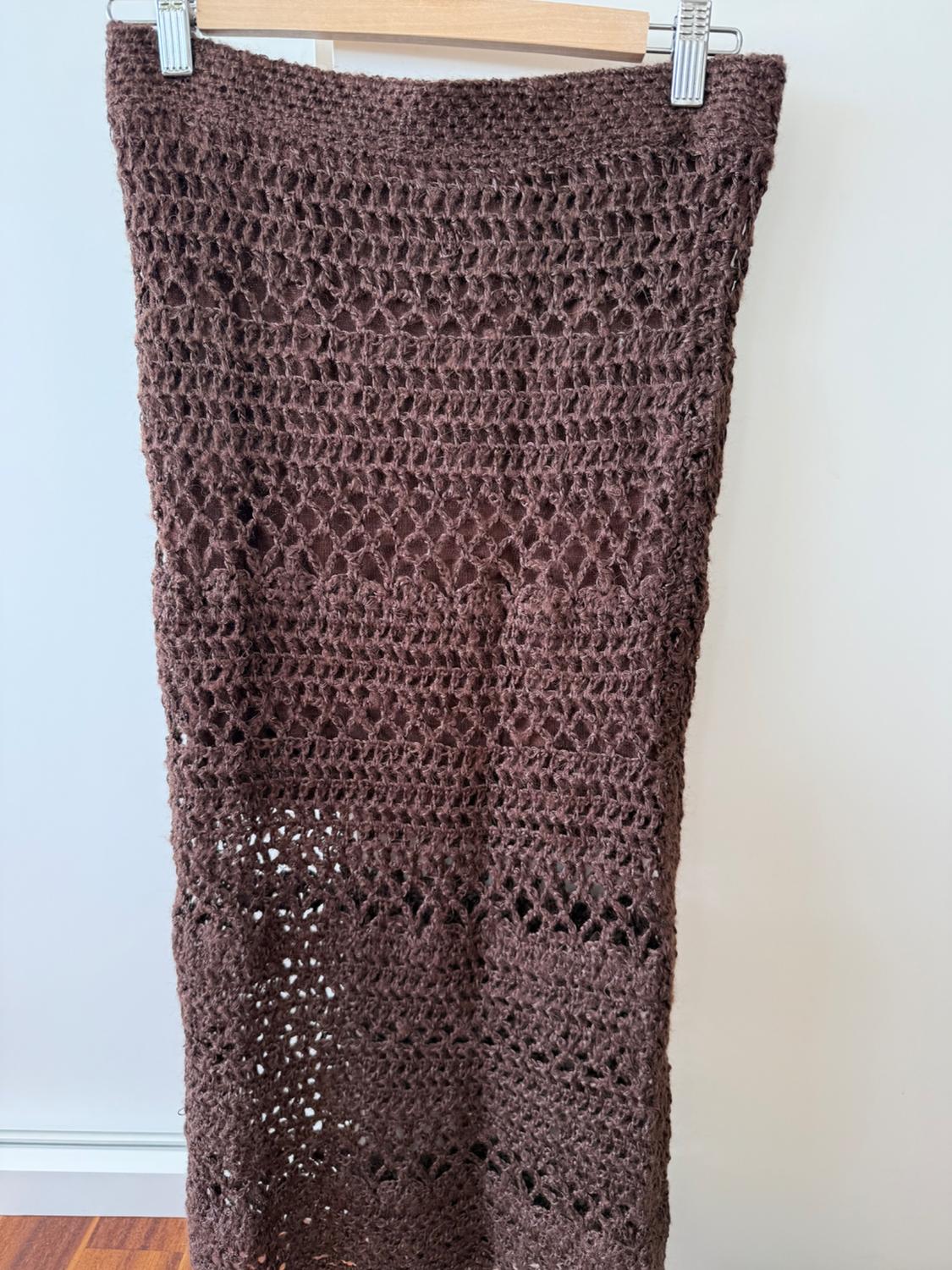 Brown crochet skirt hanging on a wooden hanger, with an open-pattern design and knitted texture, against a white background.