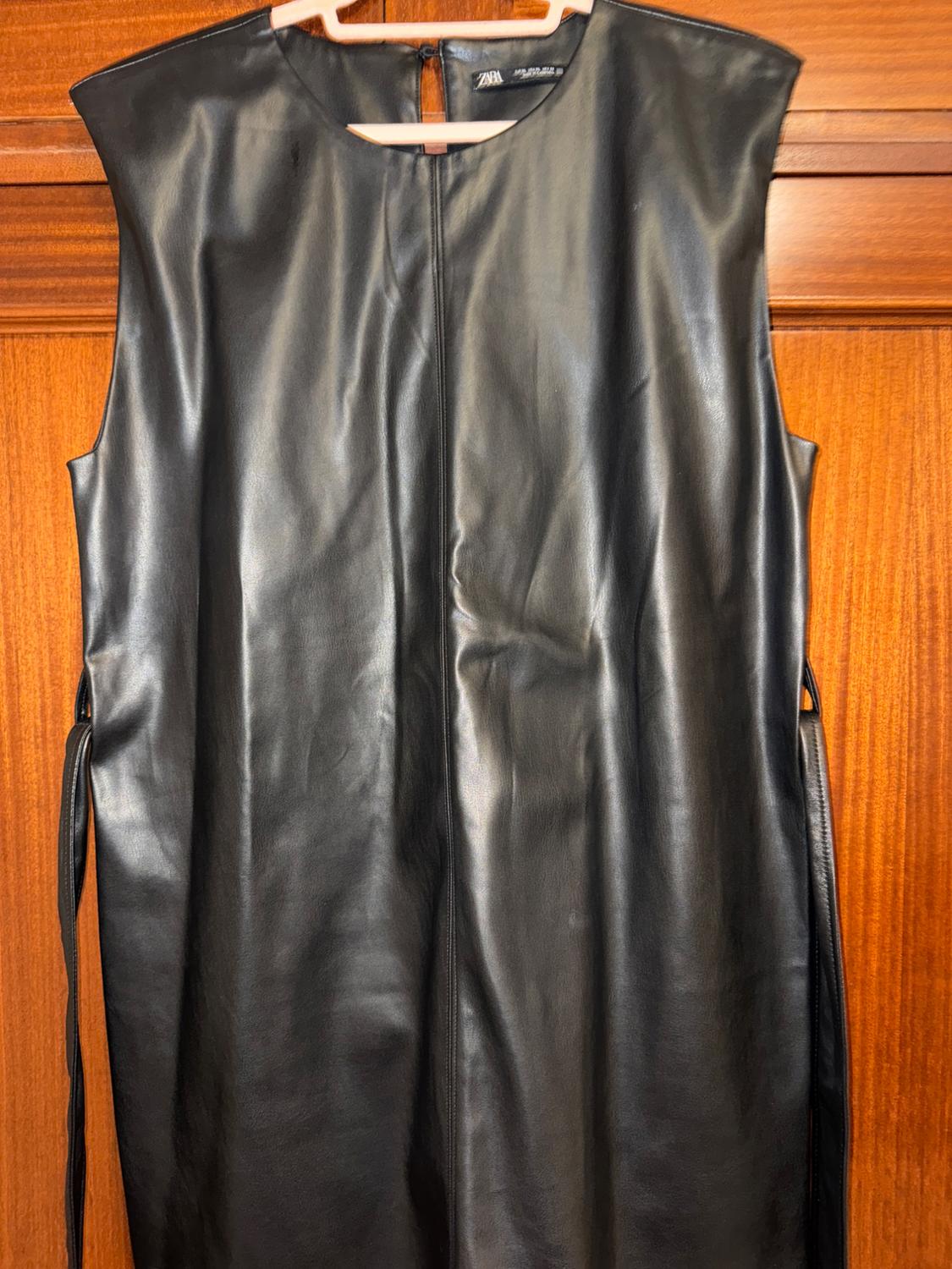 Sleeveless black dress made of leather-look material, hanging on a wooden hanger against a brown wooden background.
