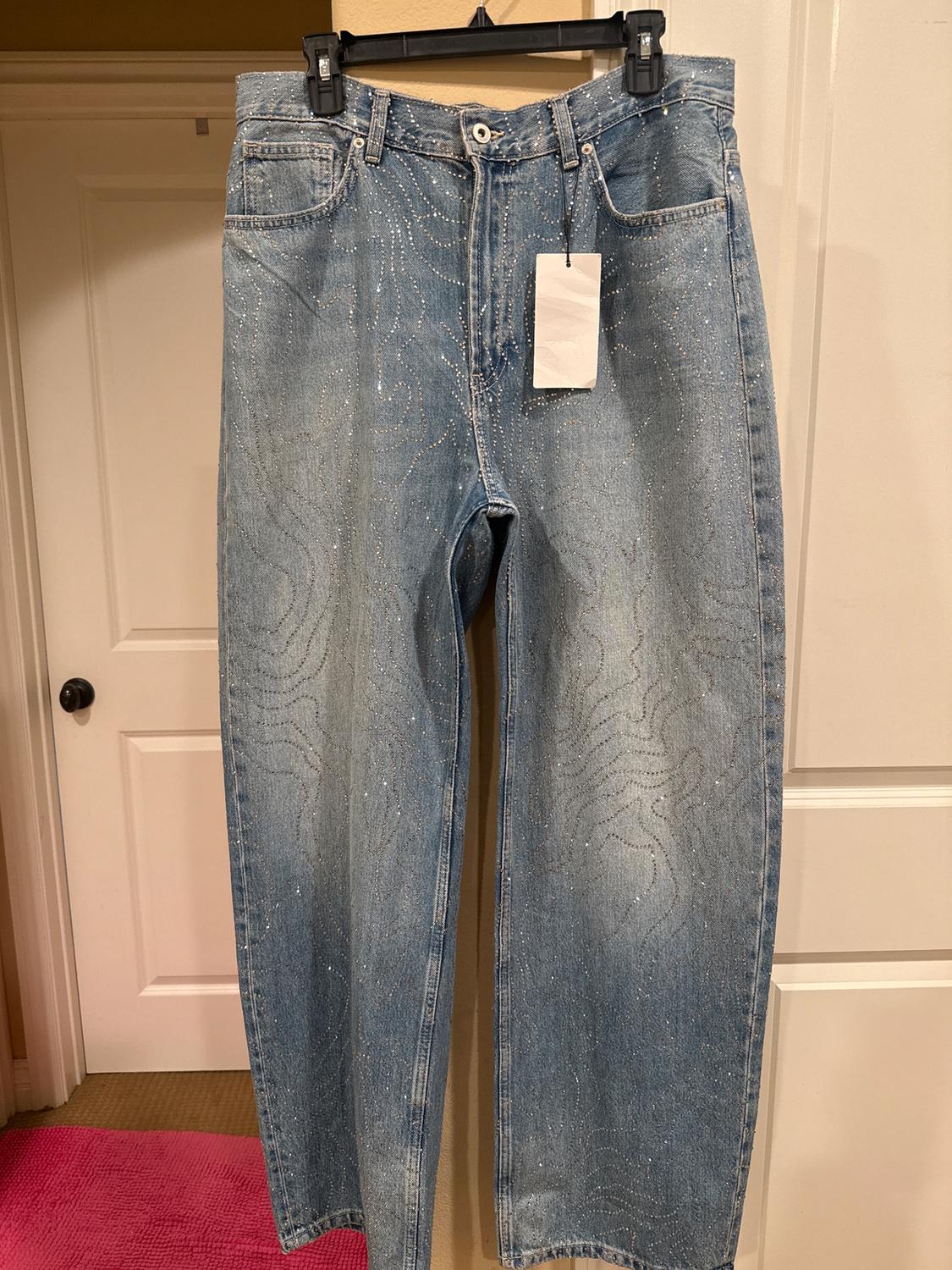 Blue jeans with glitter details hanging on a hanger. White tag visible. White doors and pink carpet in the background.