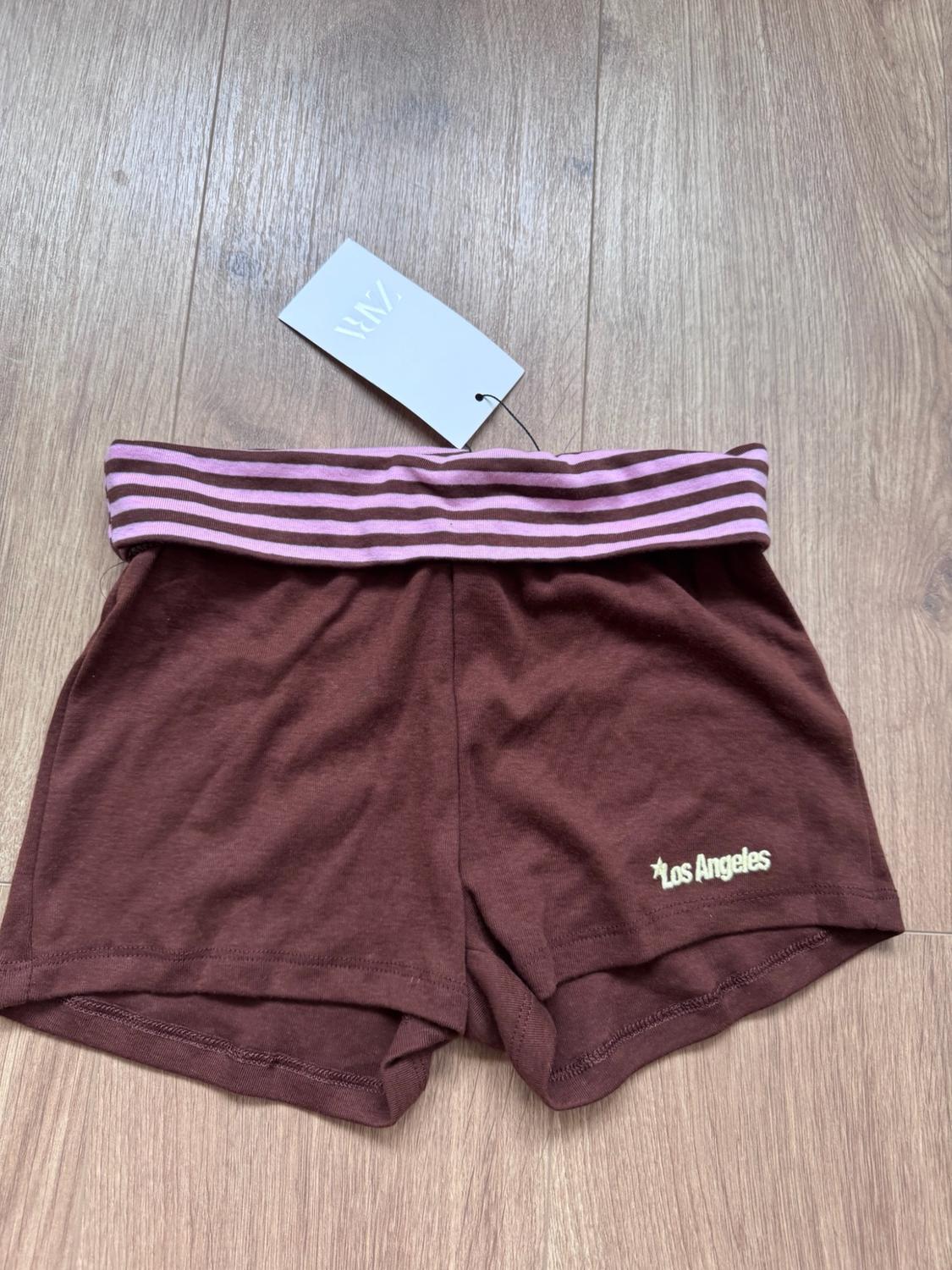 Brown shorts with a pink striped waistband on a wooden floor. It features a ZARA label and the text ‘Los Angeles’.
