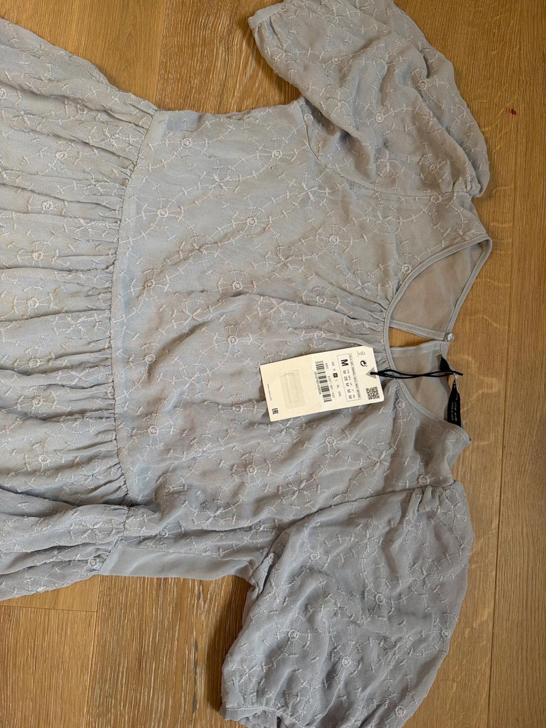 Light grey dress with puff sleeves and floral embroidery on a wooden background. Visible label with barcode.