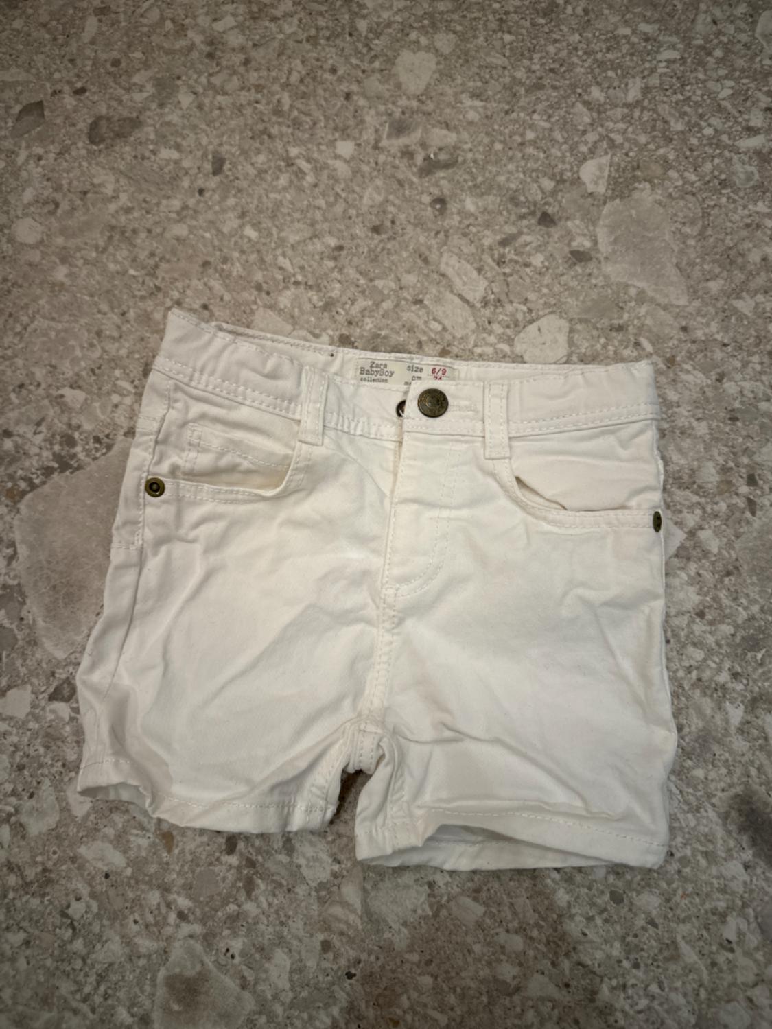 White baby shorts with pockets and a front button, on a grey stone floor. Visible label: "Zara Babyboy, size 6/9".